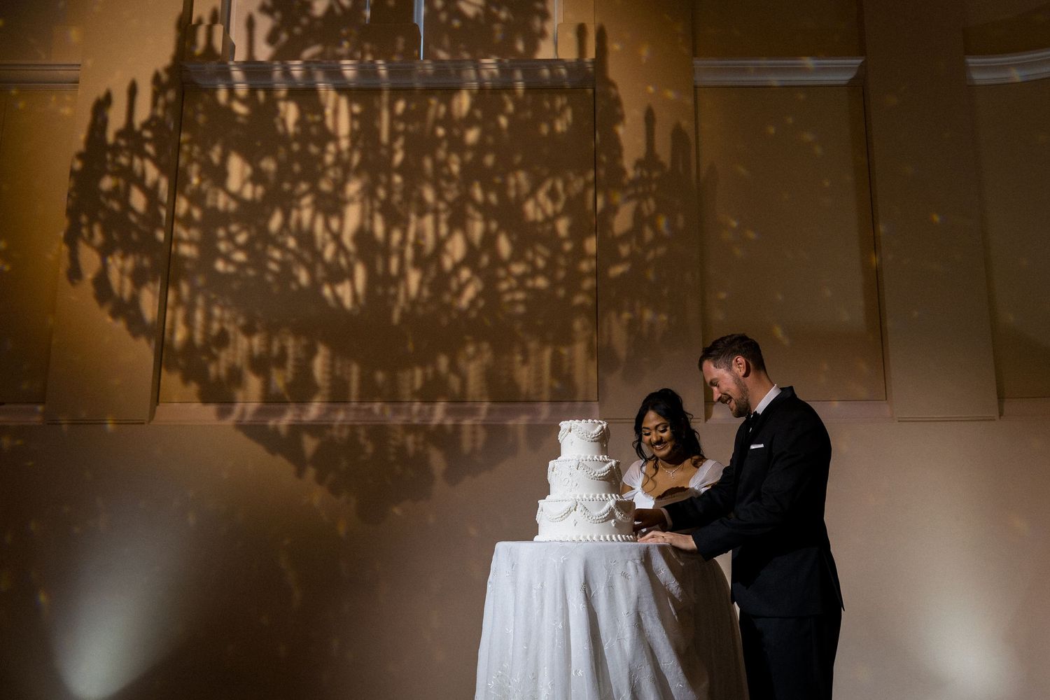 Bride and groom cut white wedding cake under dramatic chandelier shadow at elegant reception. at Montgomery Hall