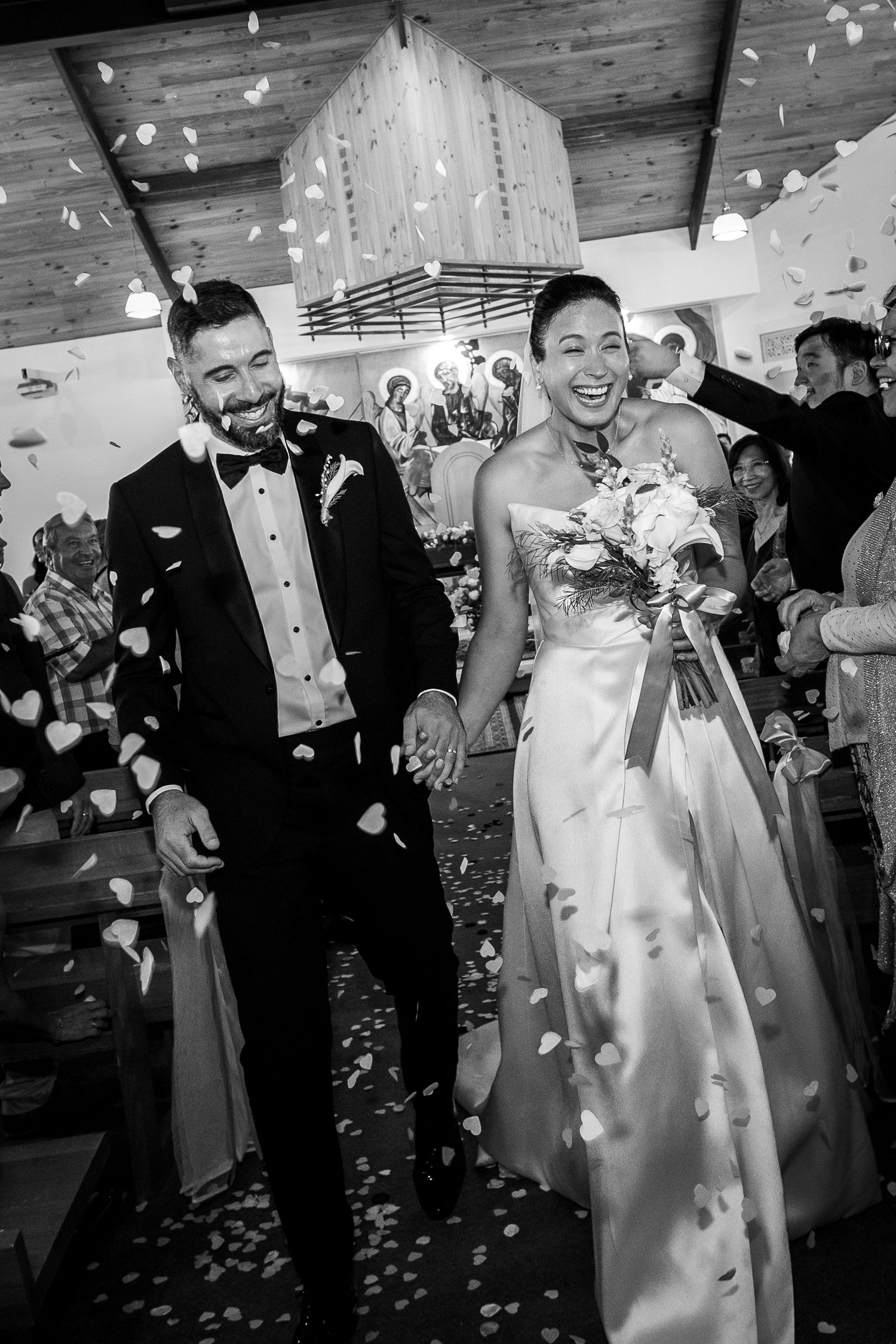 Black and white photo of a joyful bride and groom walking down the aisle as flower petals rain down.