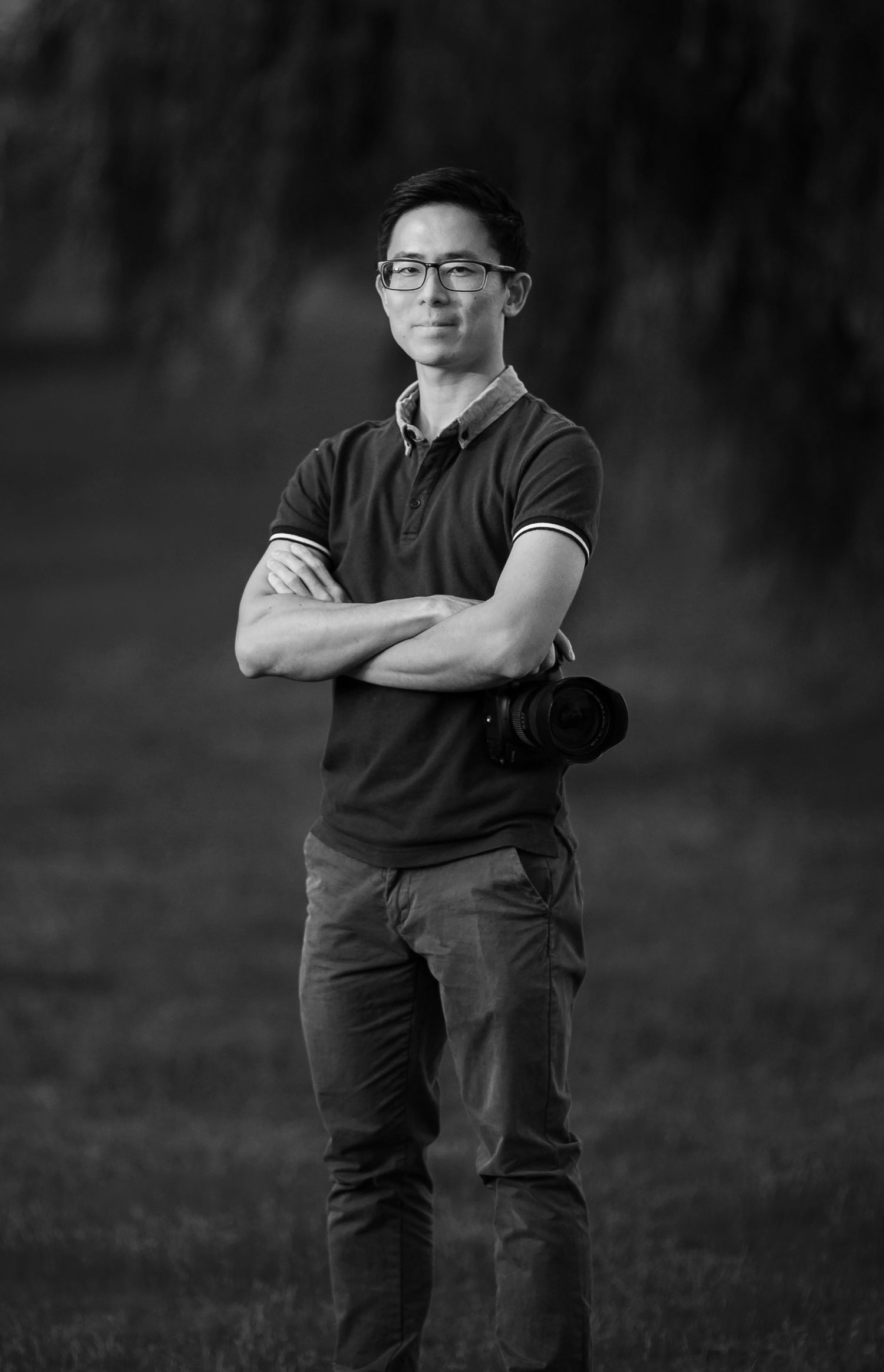 Professional photographer stands with arms crossed holding camera in a confident black and white portrait.