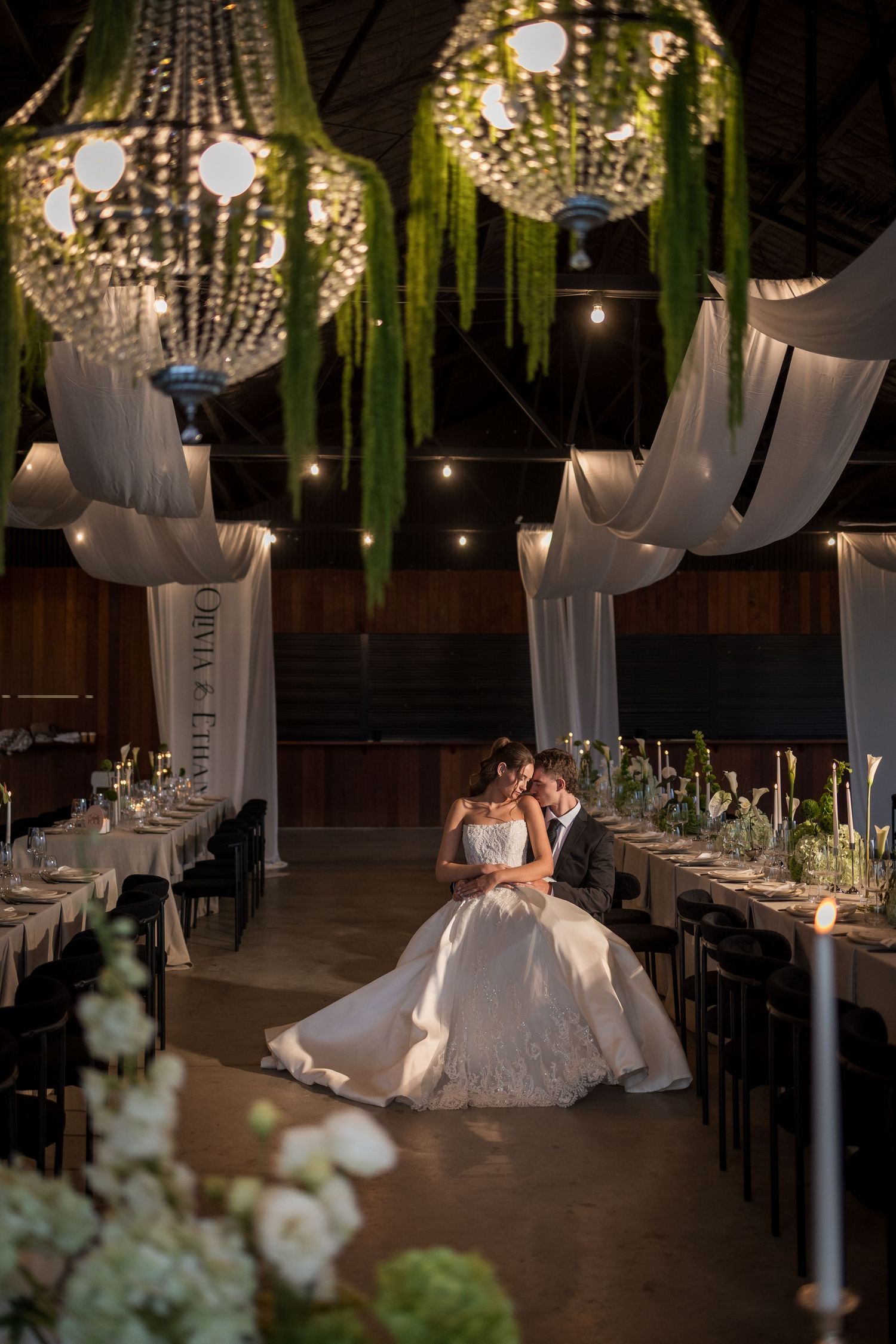 Bride and groom share a romantic moment in an elegantly decorated wedding reception hall with crystal chandeliers and greenery.