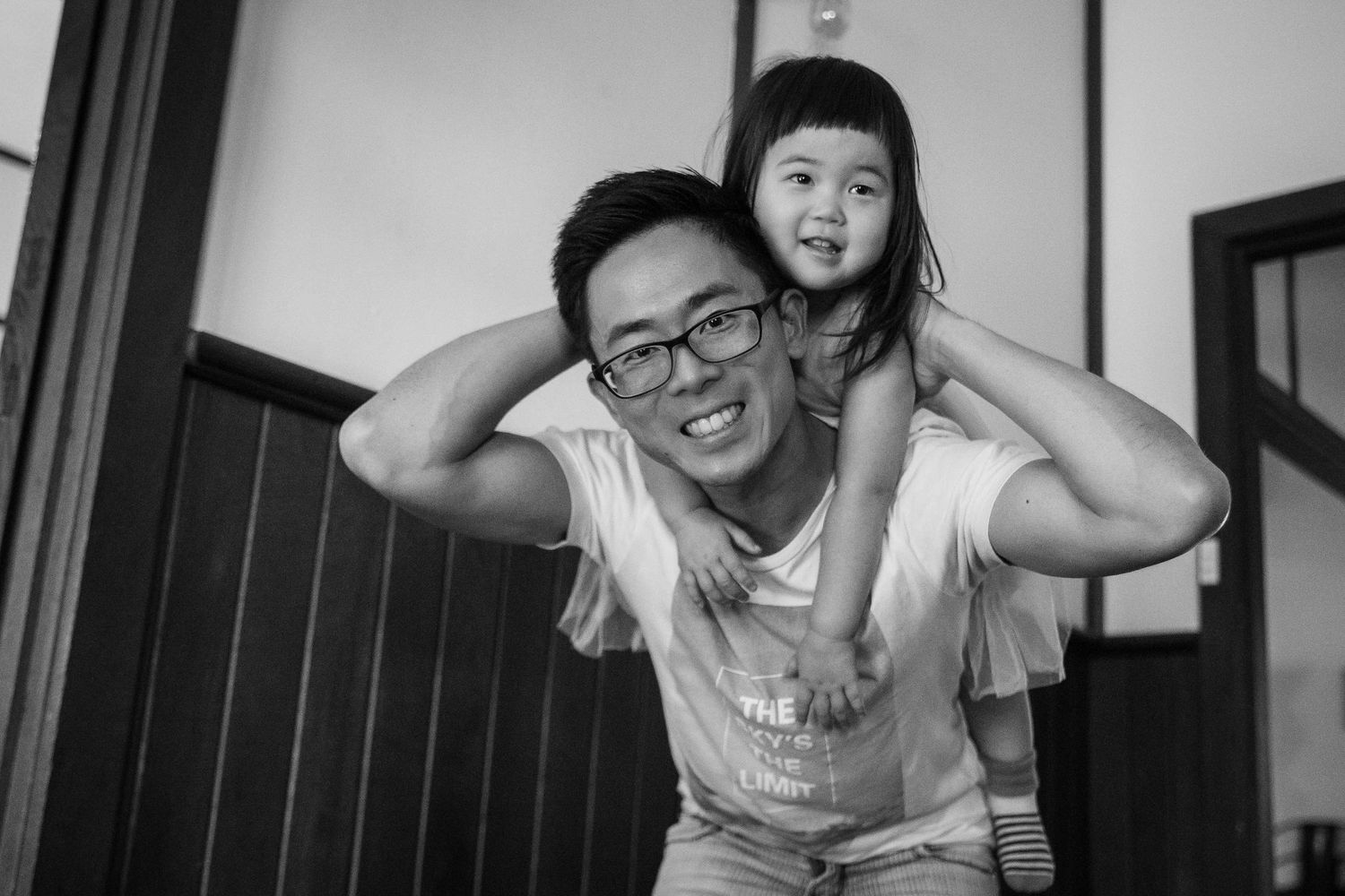 Man with glasses smiling while giving a young girl a piggyback ride in a black and white photo.