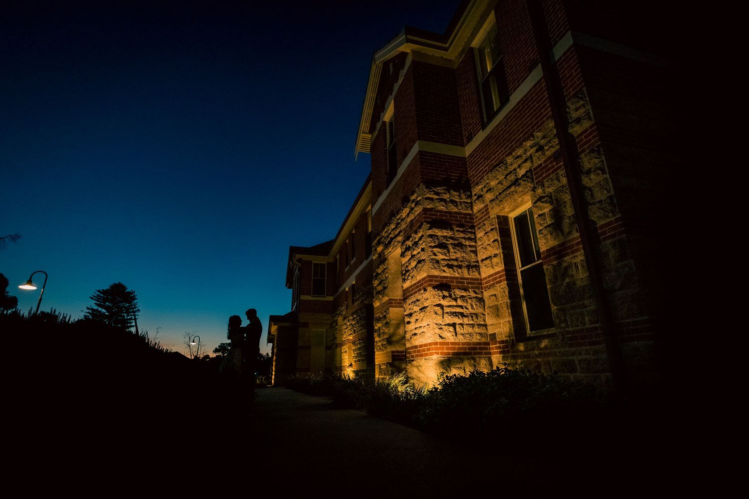 Couple silhouetted against illuminated historic stone building at dusk with deep blue sky.