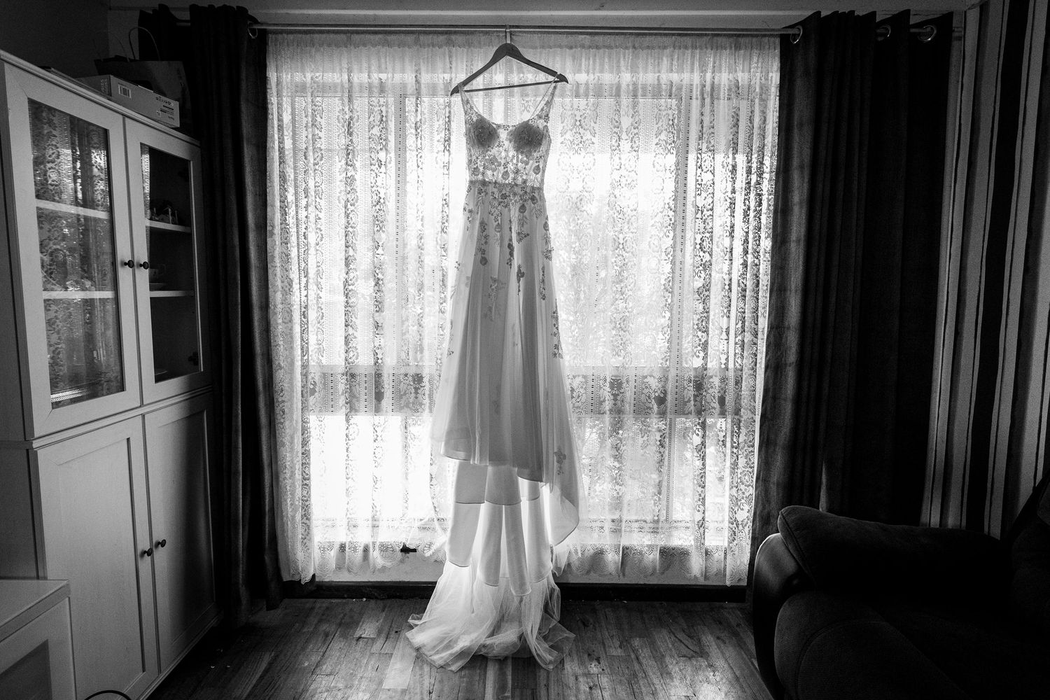 Wedding dress hangs in silhouette against bright window with lace curtains.