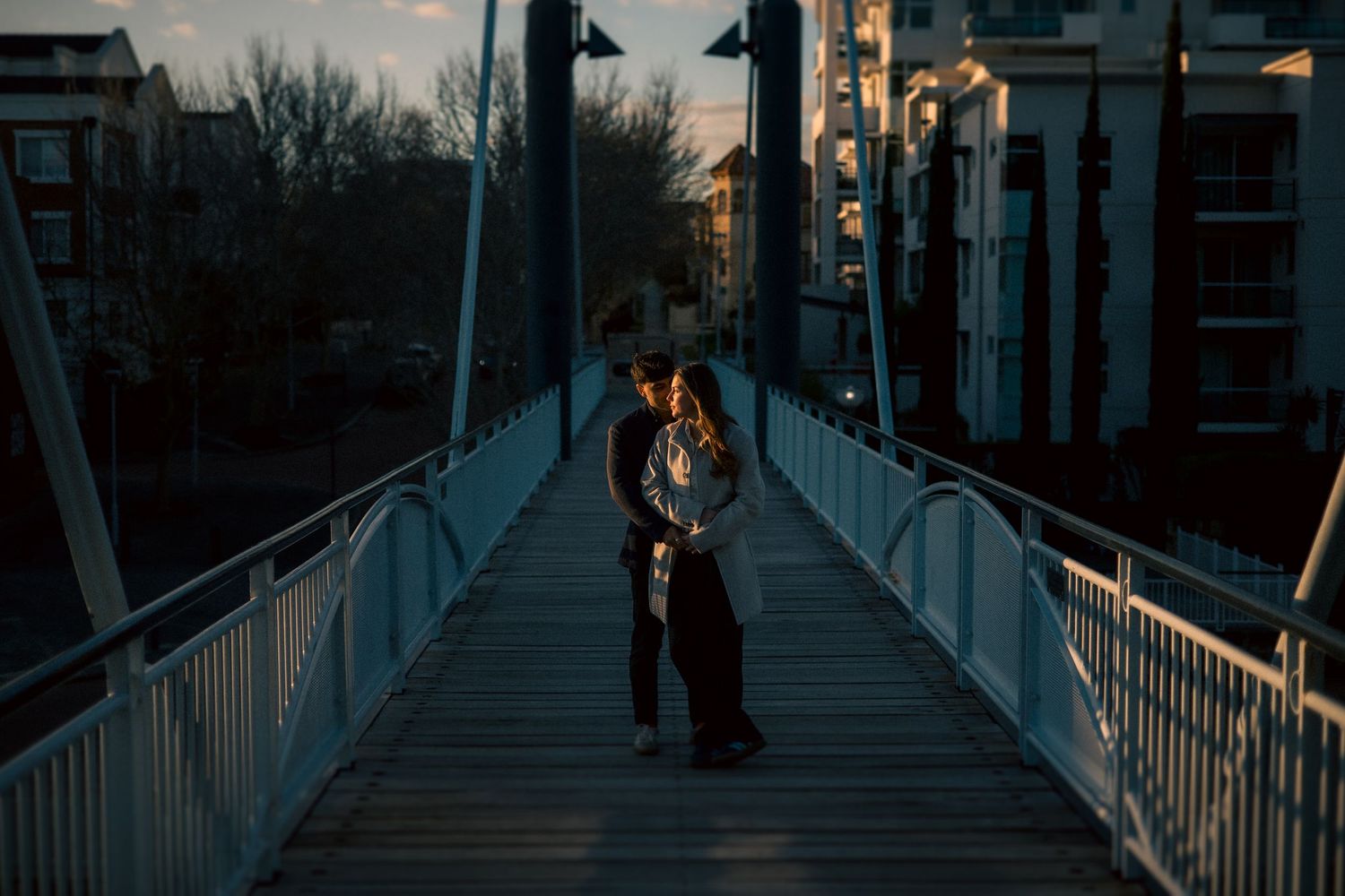 Couple sharing a romantic embrace on a pedestrian bridge at dusk with city buildings in the background.