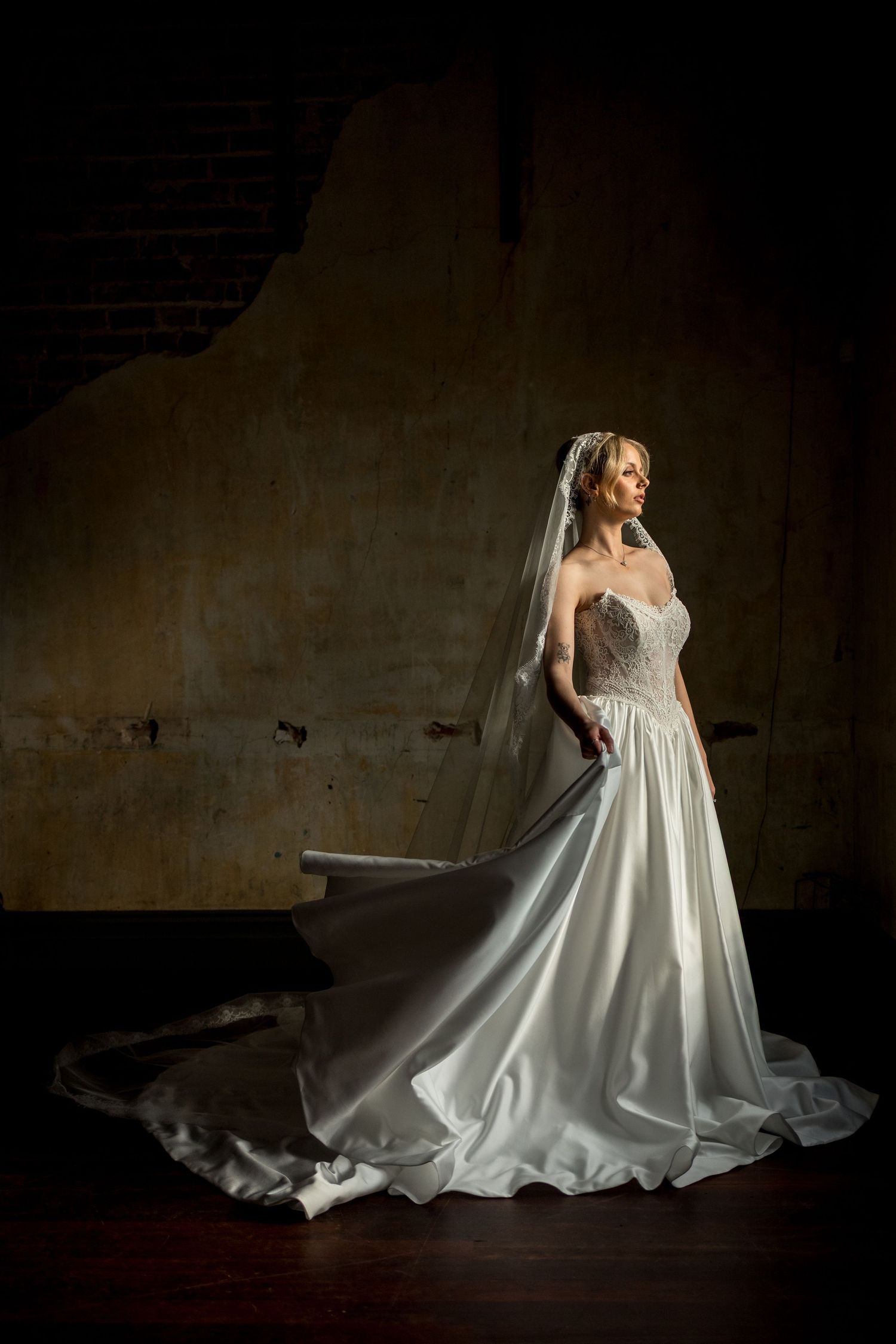 Bride in white ballgown with long veil stands in dramatic light against stone wall.