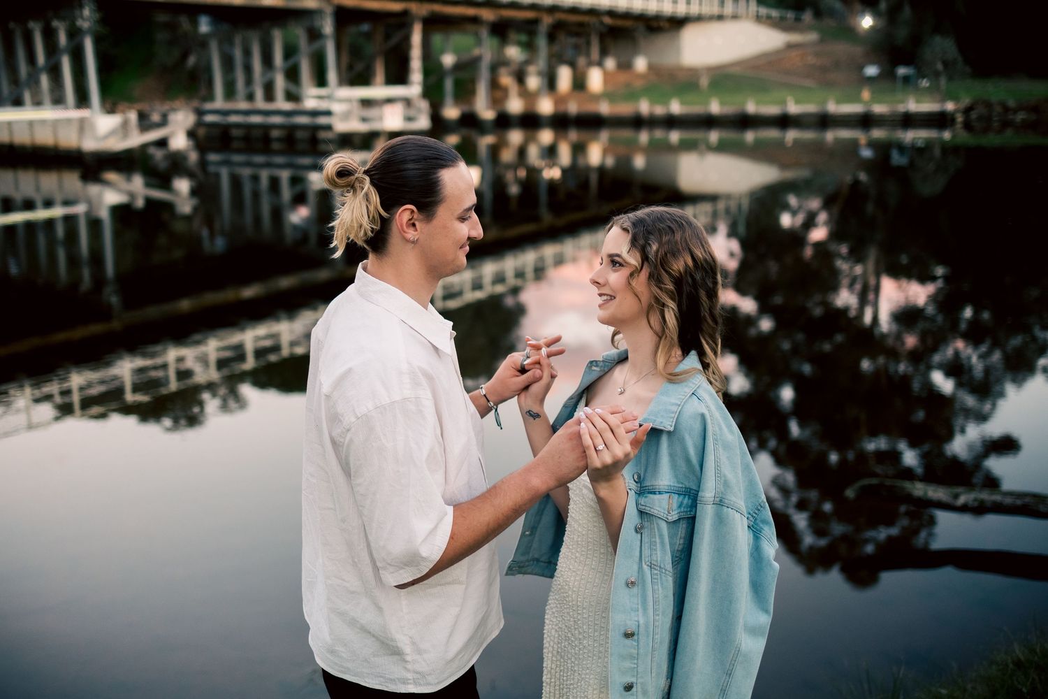 A couple holds hands and smiles at each other by a reflective river near a bridge at dusk. at Guildford, Perth