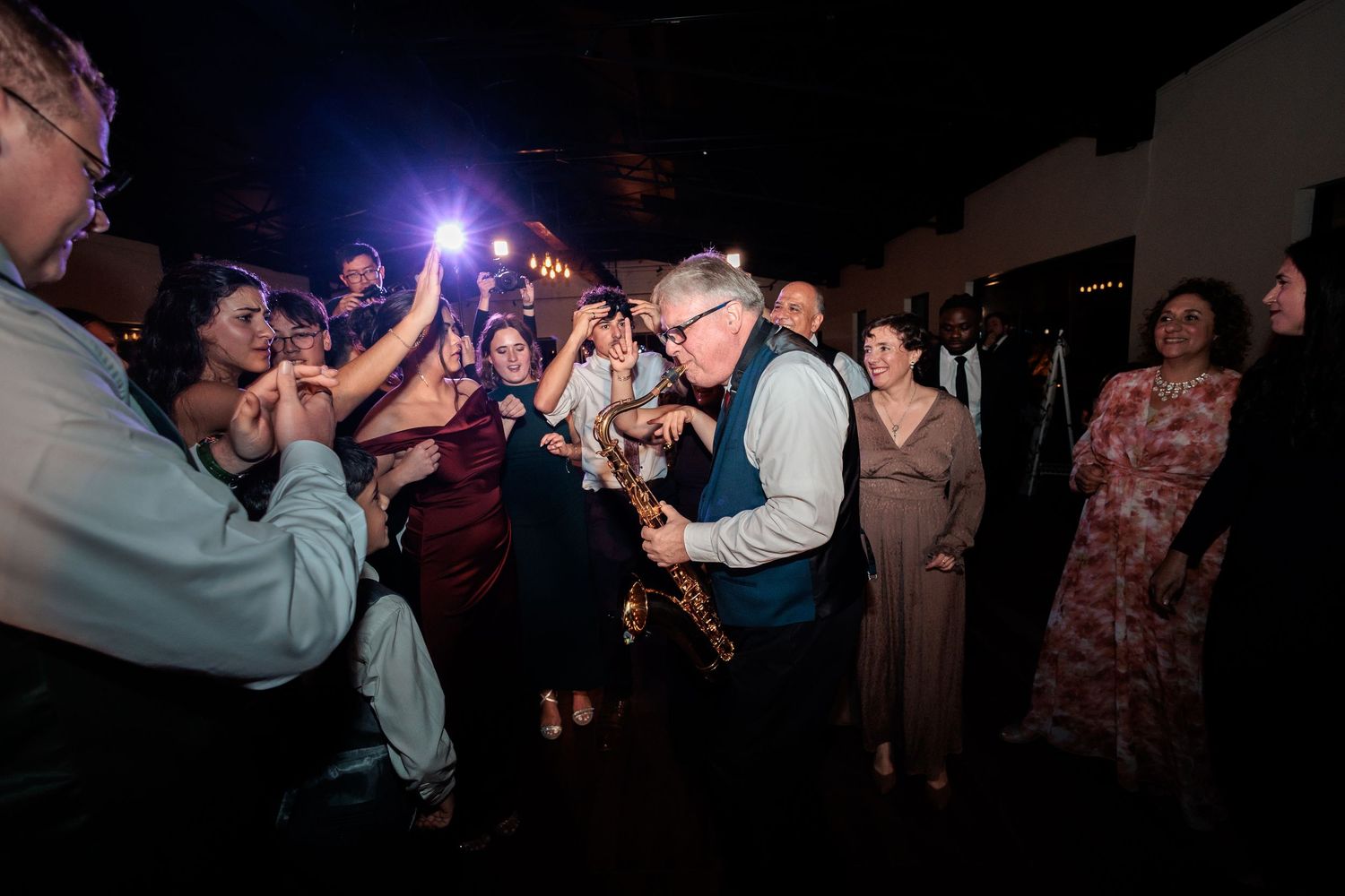 A saxophonist performs for guests on a wedding dance floor while attendees cheer and photograph the lively celebration.