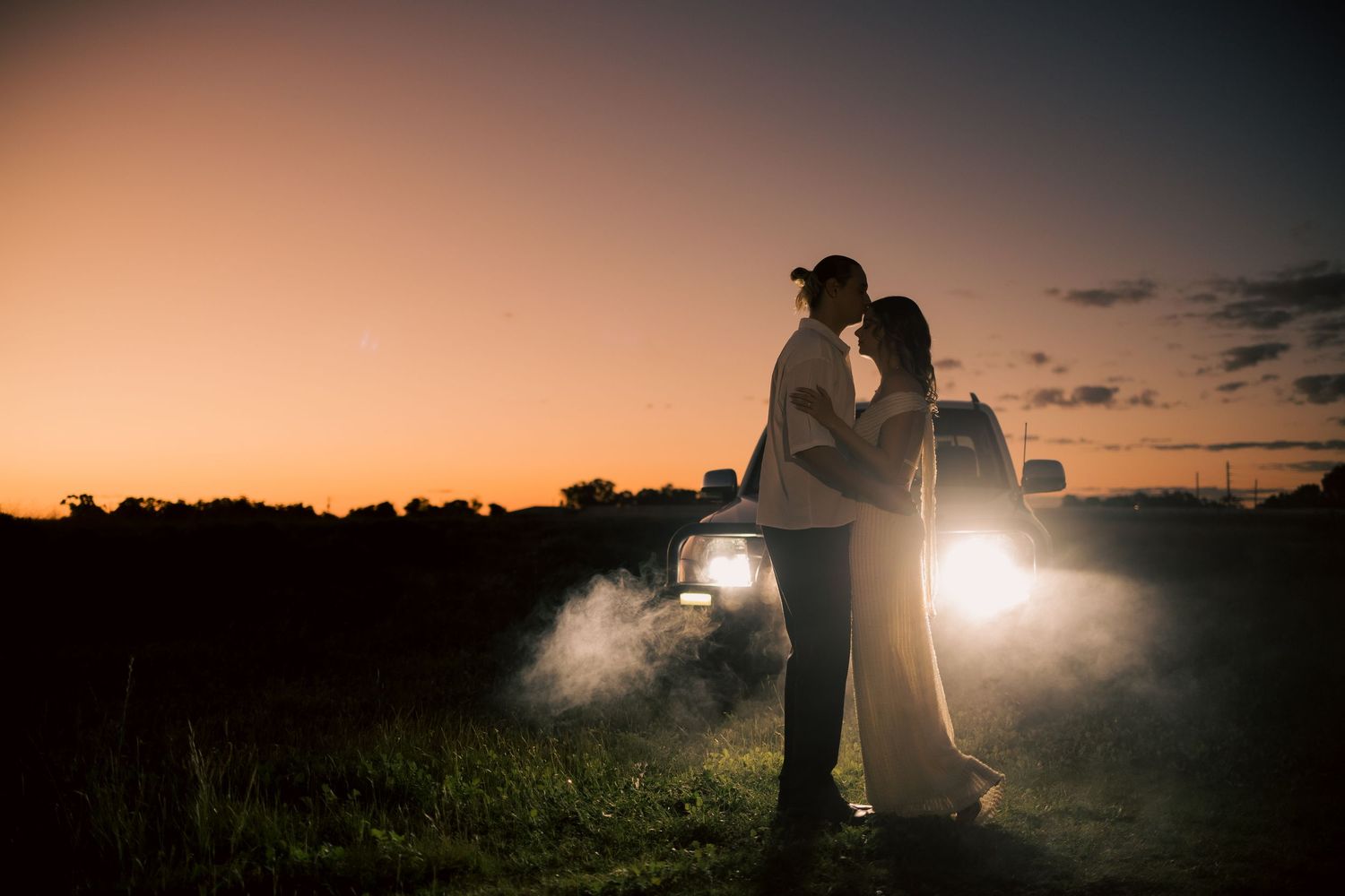 Romantic couple silhouetted against a glowing sunset, standing in front of car headlights in an open field. at Guildford, Perth