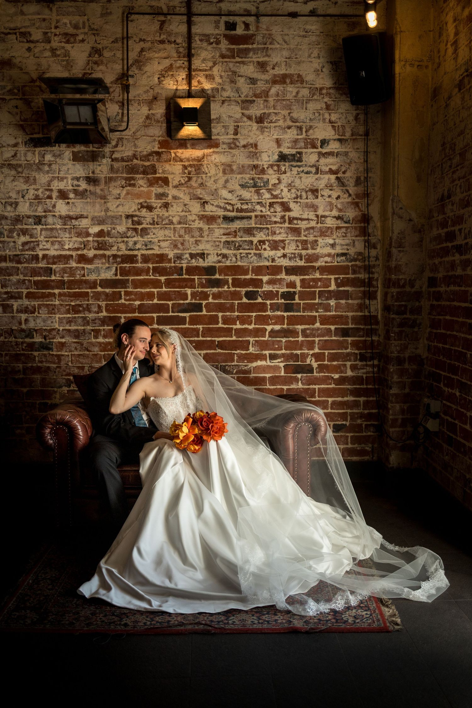 Bride and groom share intimate moment on leather chair against rustic brick wall, bridal veil flowing with orange bouquet.