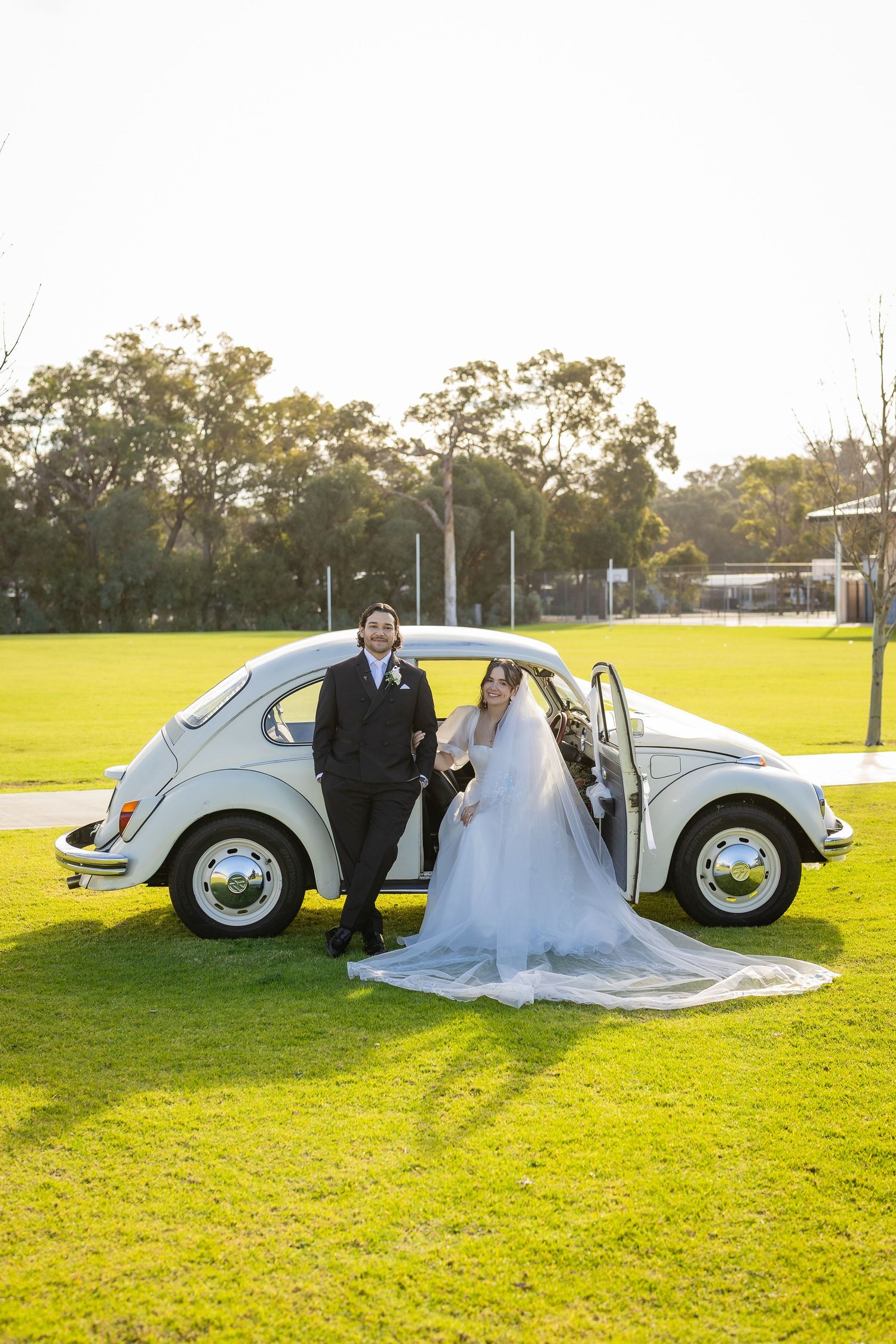 Bride and groom posing beside vintage white VW Beetle car on green lawn at sunset.