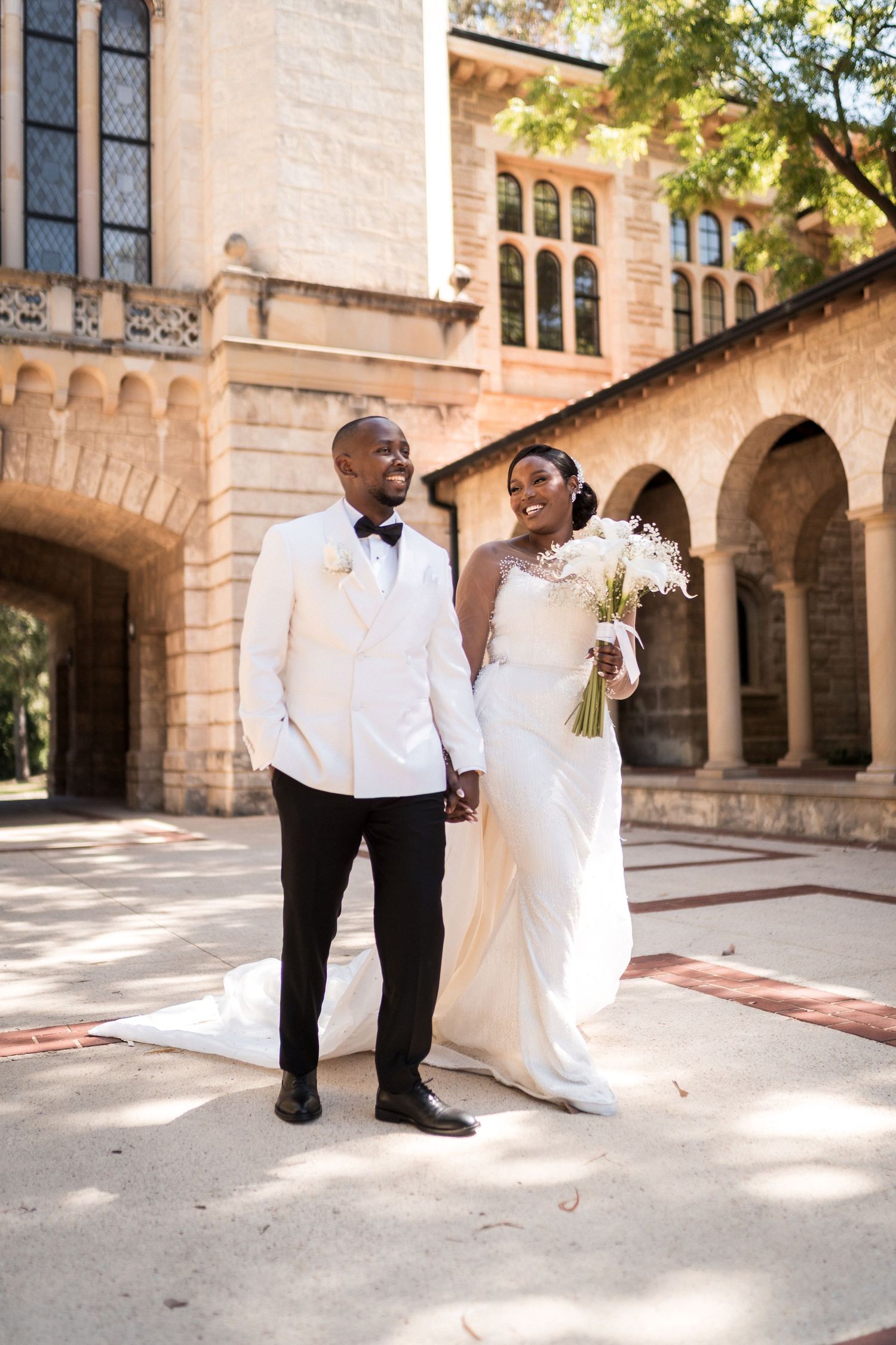 A Black bride and groom in wedding attire hold hands outside a historic stone building with arched walkways.