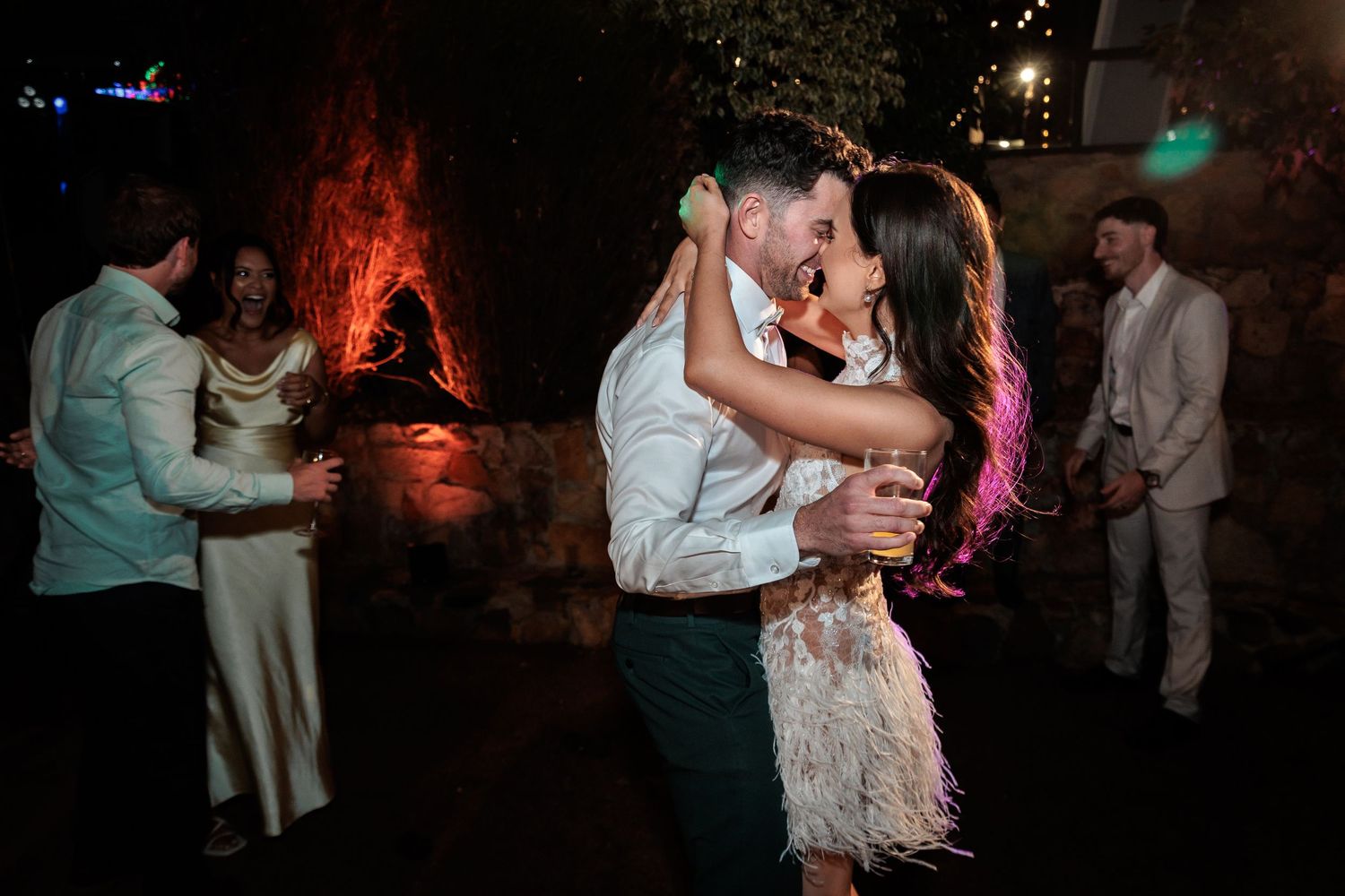 Couple shares romantic dance at wedding reception with guests celebrating in background under warm evening lights.