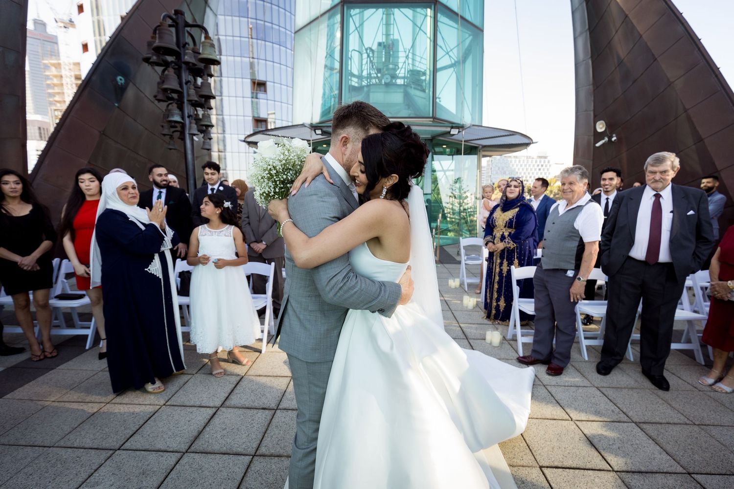 Bride and groom share first kiss at outdoor rooftop wedding ceremony with guests watching in background.