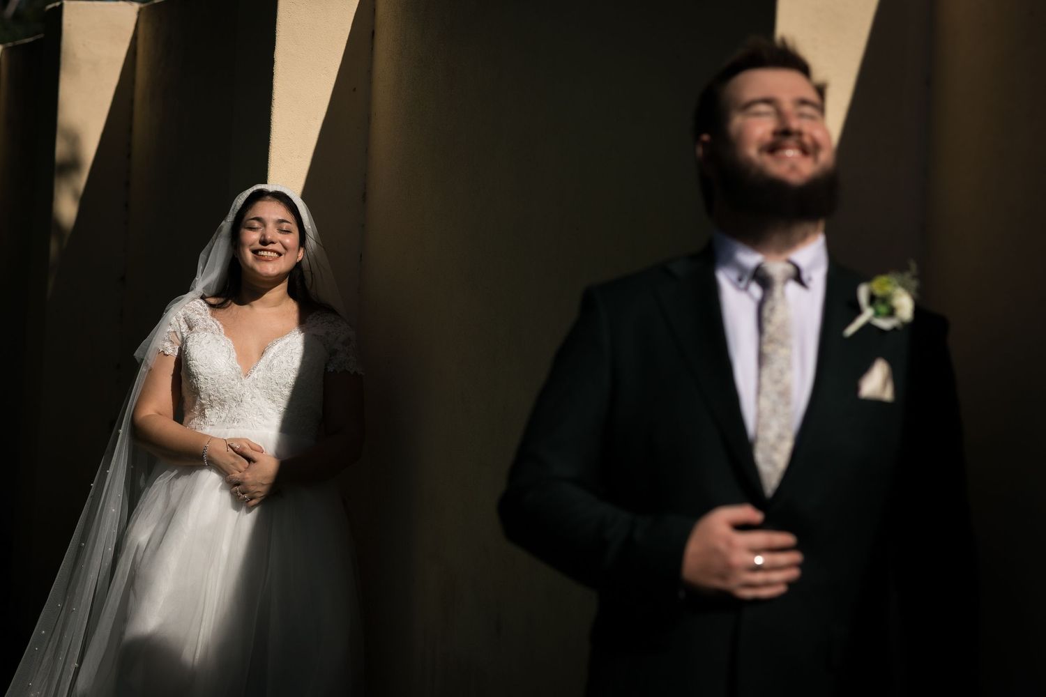 Bride in white gown and veil smiles softly while groom in dark suit stands in foreground during wedding portrait.