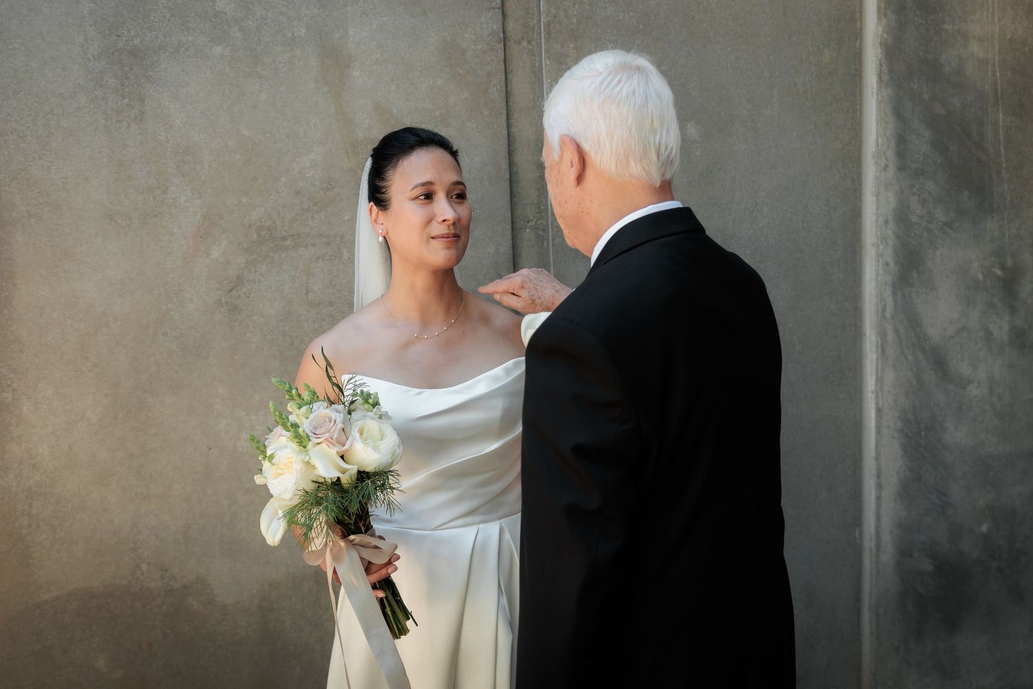 Bride in white gown holding bouquet smiles at older gentleman in tuxedo against gray wall.