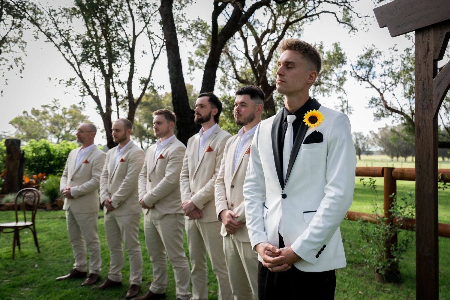 Groomsmen in cream suits lined up outdoors under trees for formal wedding photos.