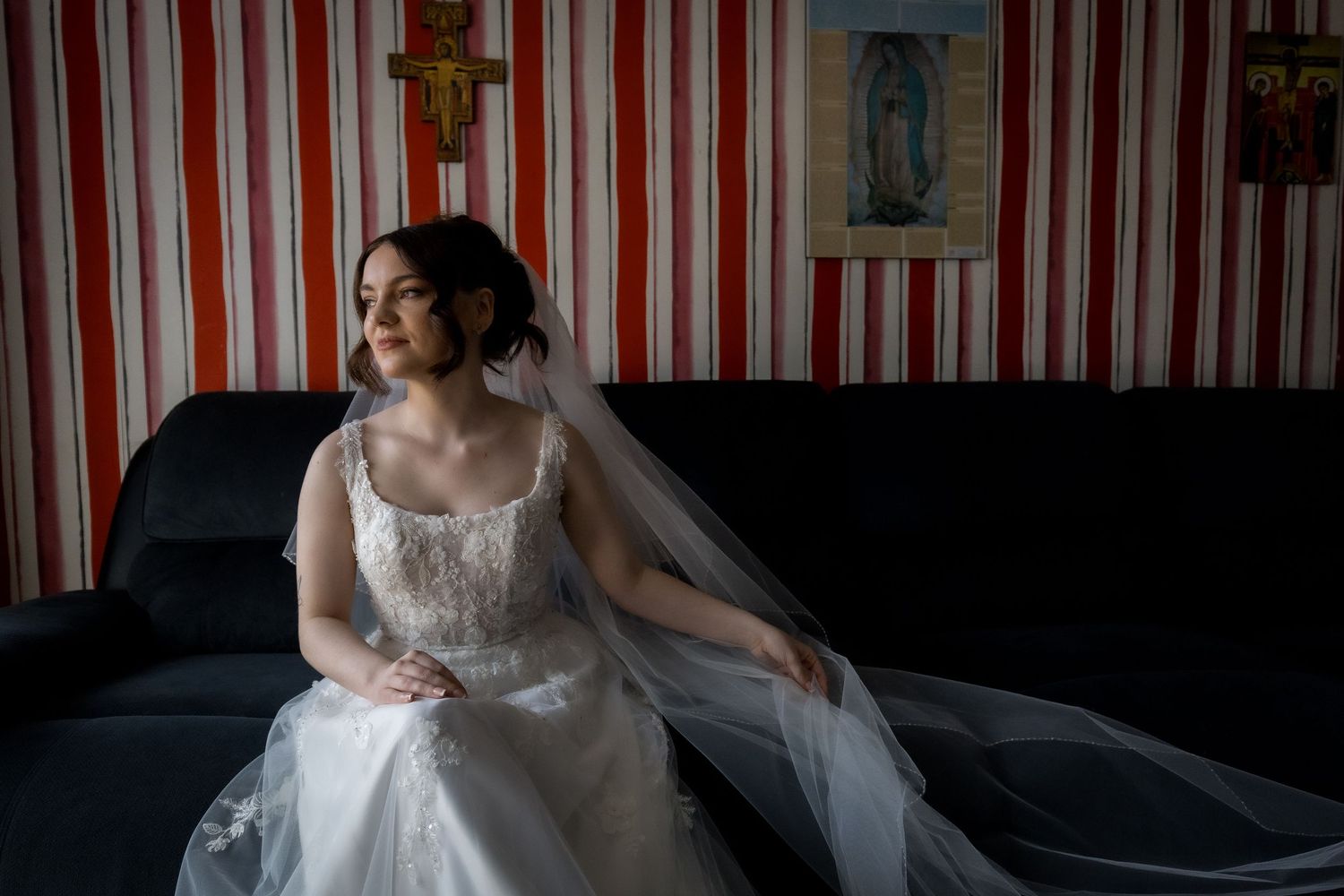 Bride in white dress sits on dark couch with striped wallpaper background.