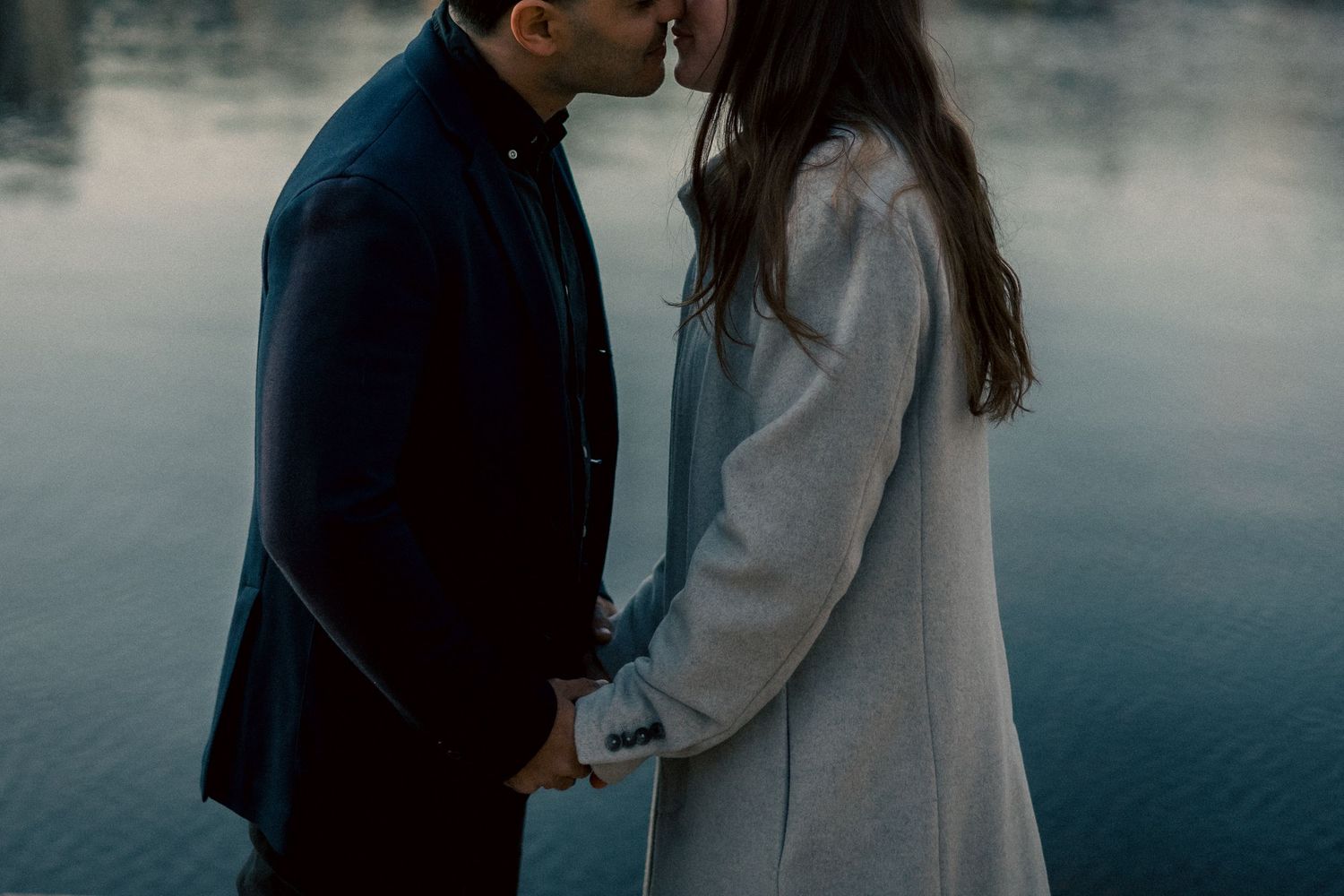 Couple shares intimate kiss while holding hands beside a calm reflective lake.