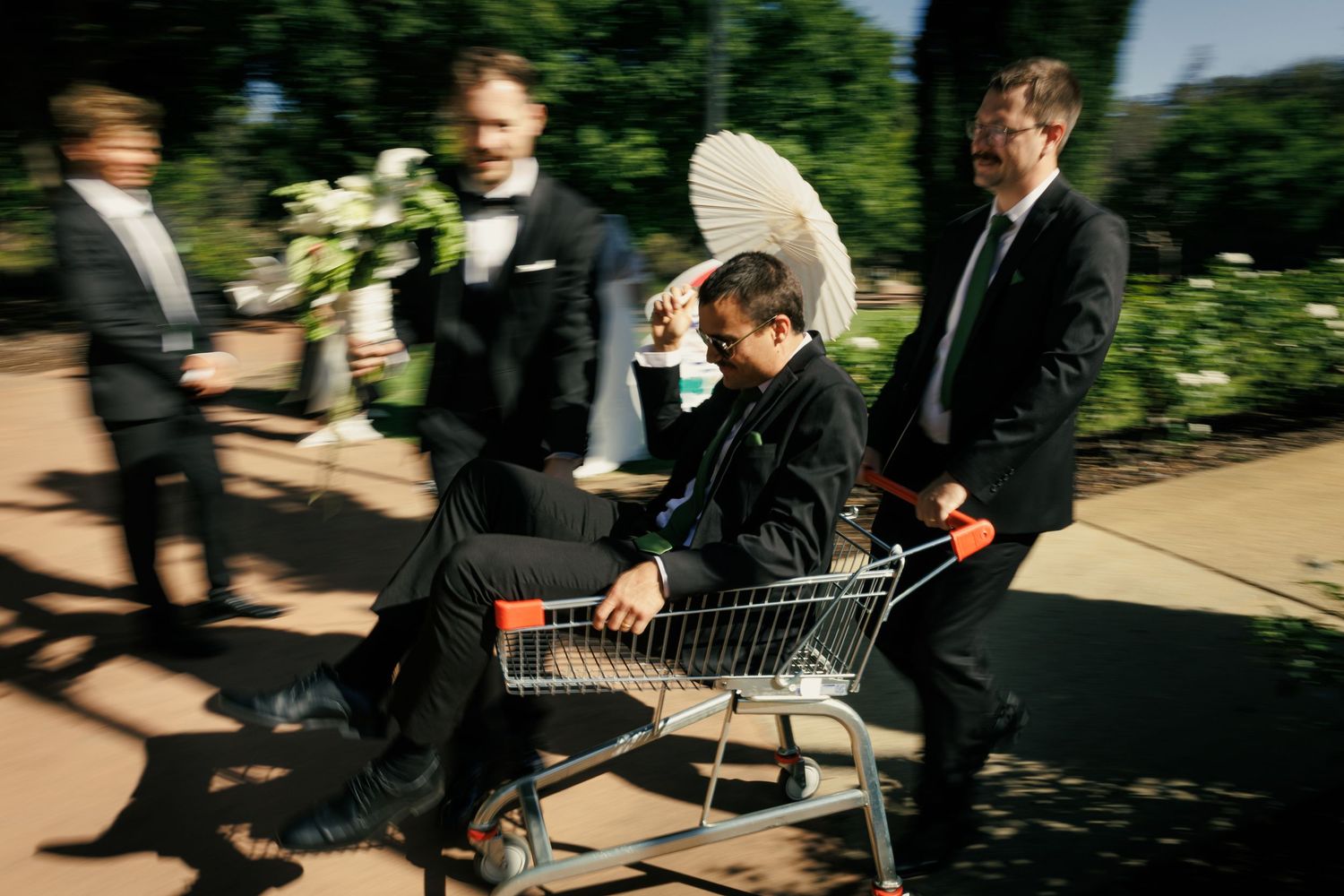 Groom rides in shopping cart pushed by groomsmen in tuxedos at outdoor wedding celebration.