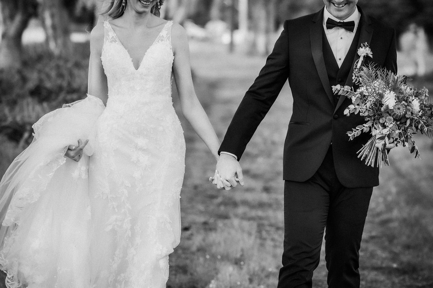 Black and white photo of a bride and groom holding hands, walking together on their wedding day.