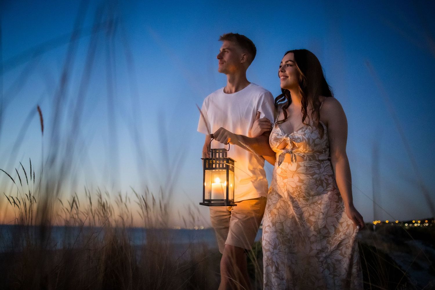 Couple holding a glowing lantern at sunset on a beach, romantic evening scene with tall grass and twilight sky.