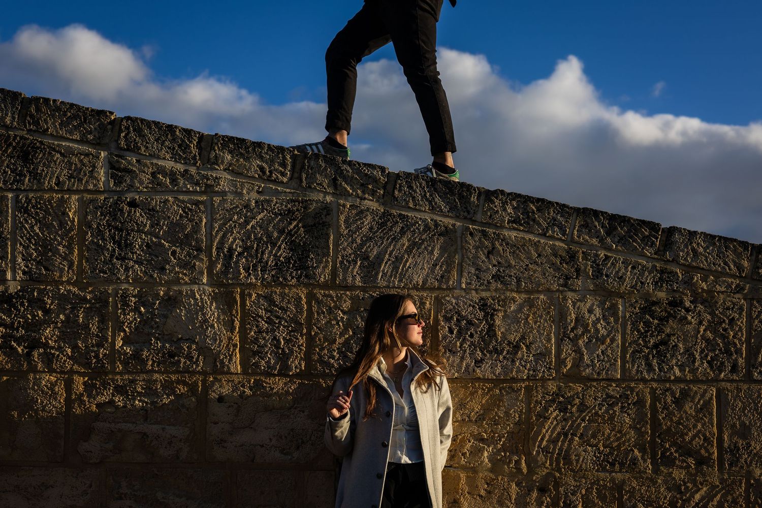 Two figures stand against a stone wall under bright blue skies and puffy white clouds.