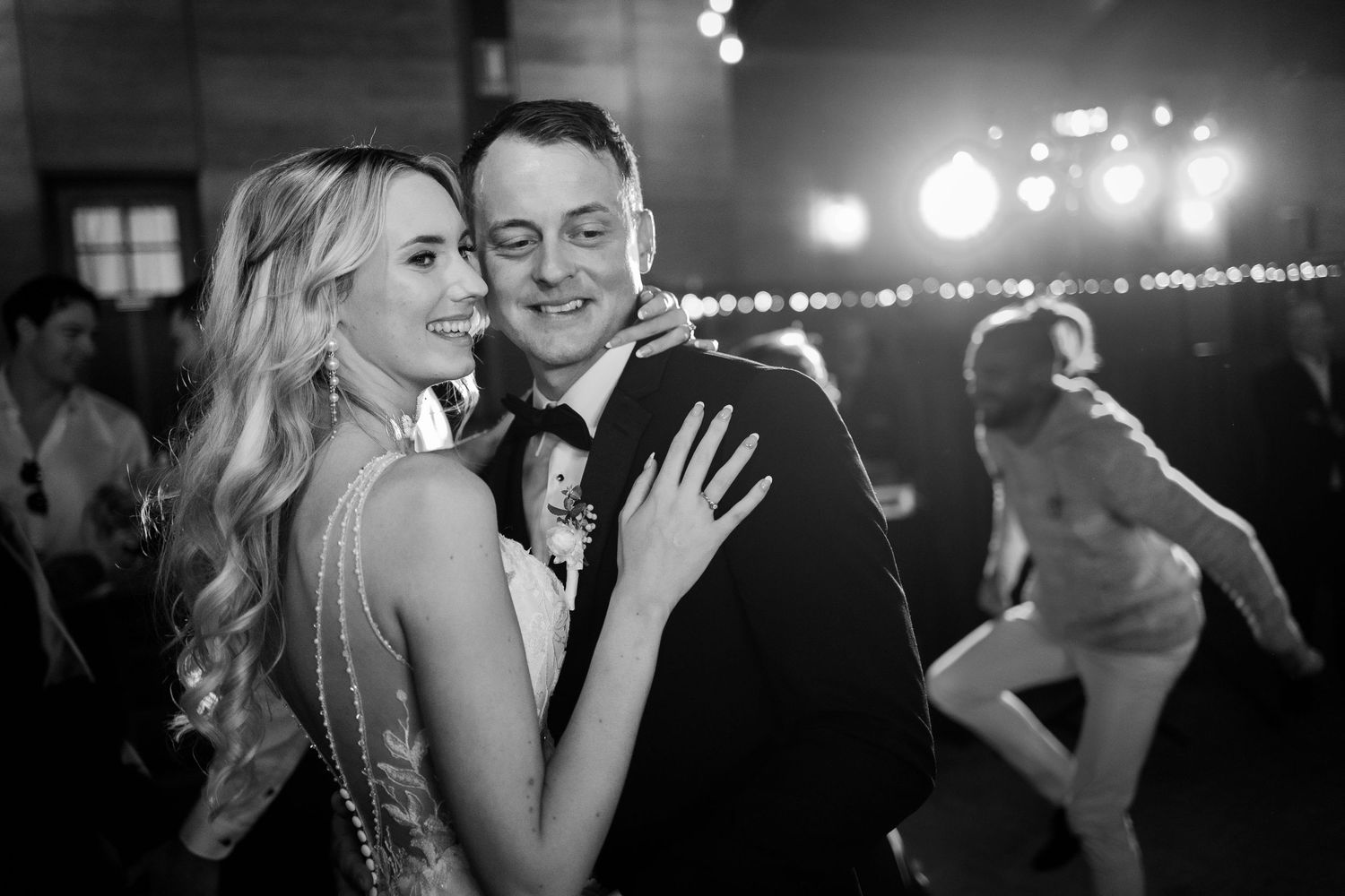 at Quarry Farm- Black and white photo of a smiling couple embracing at a wedding reception with guests dancing in the background.