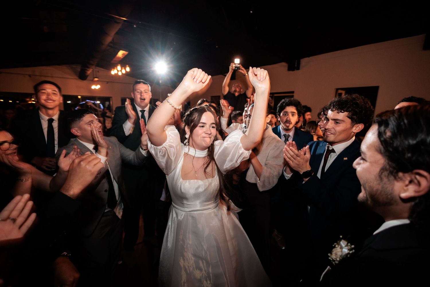Bride in white gown celebrates joyfully with hands raised surrounded by cheering wedding guests.
