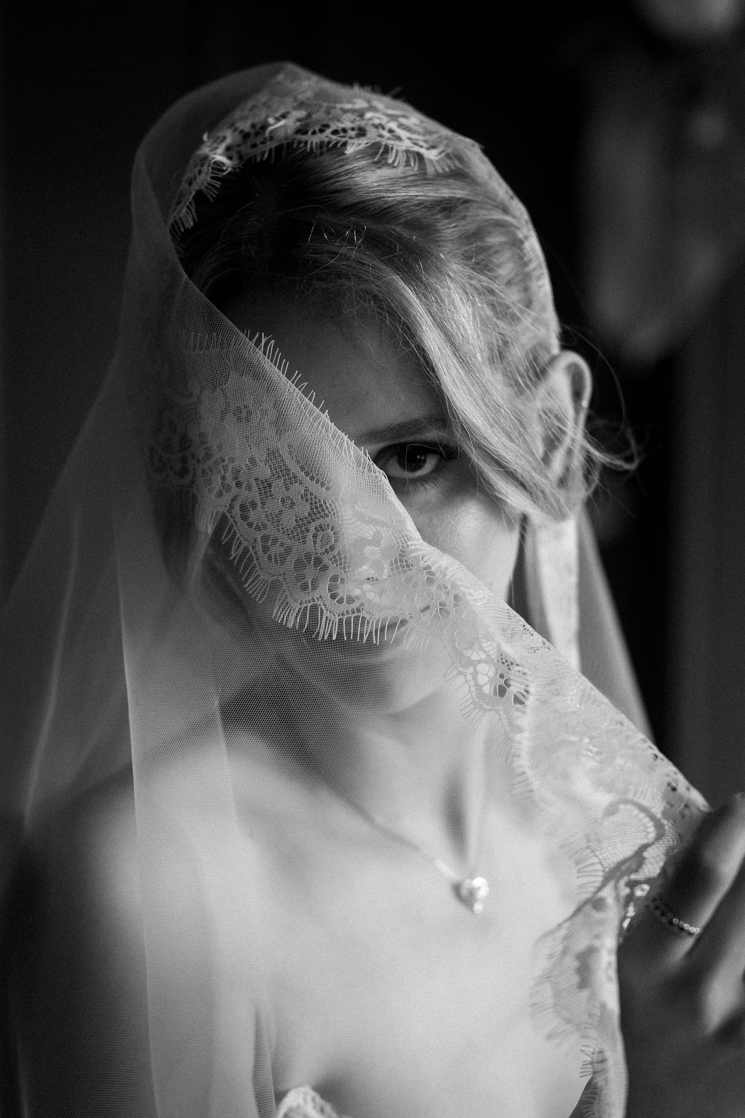Black and white portrait of a bride with lace veil draped over her face, wearing a heart pendant necklace.