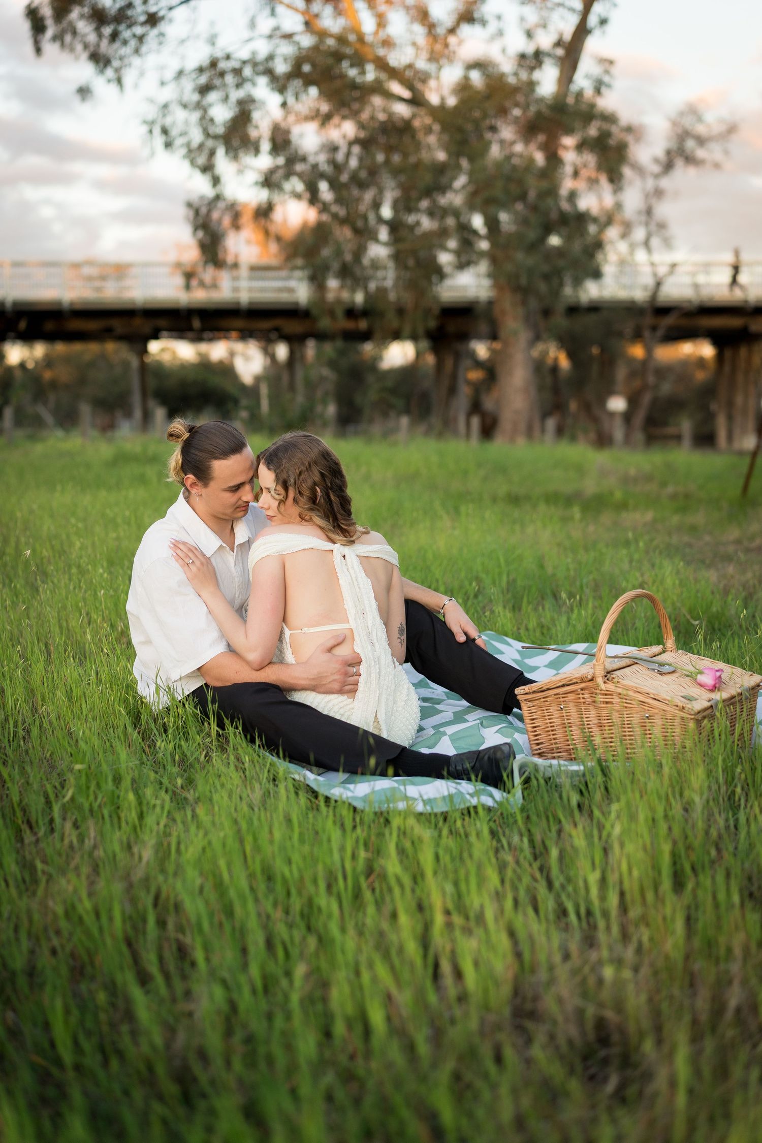 Couple sharing a romantic picnic on a green grass field at sunset, with a wicker basket and bridge in the background. at Guildford, Perth