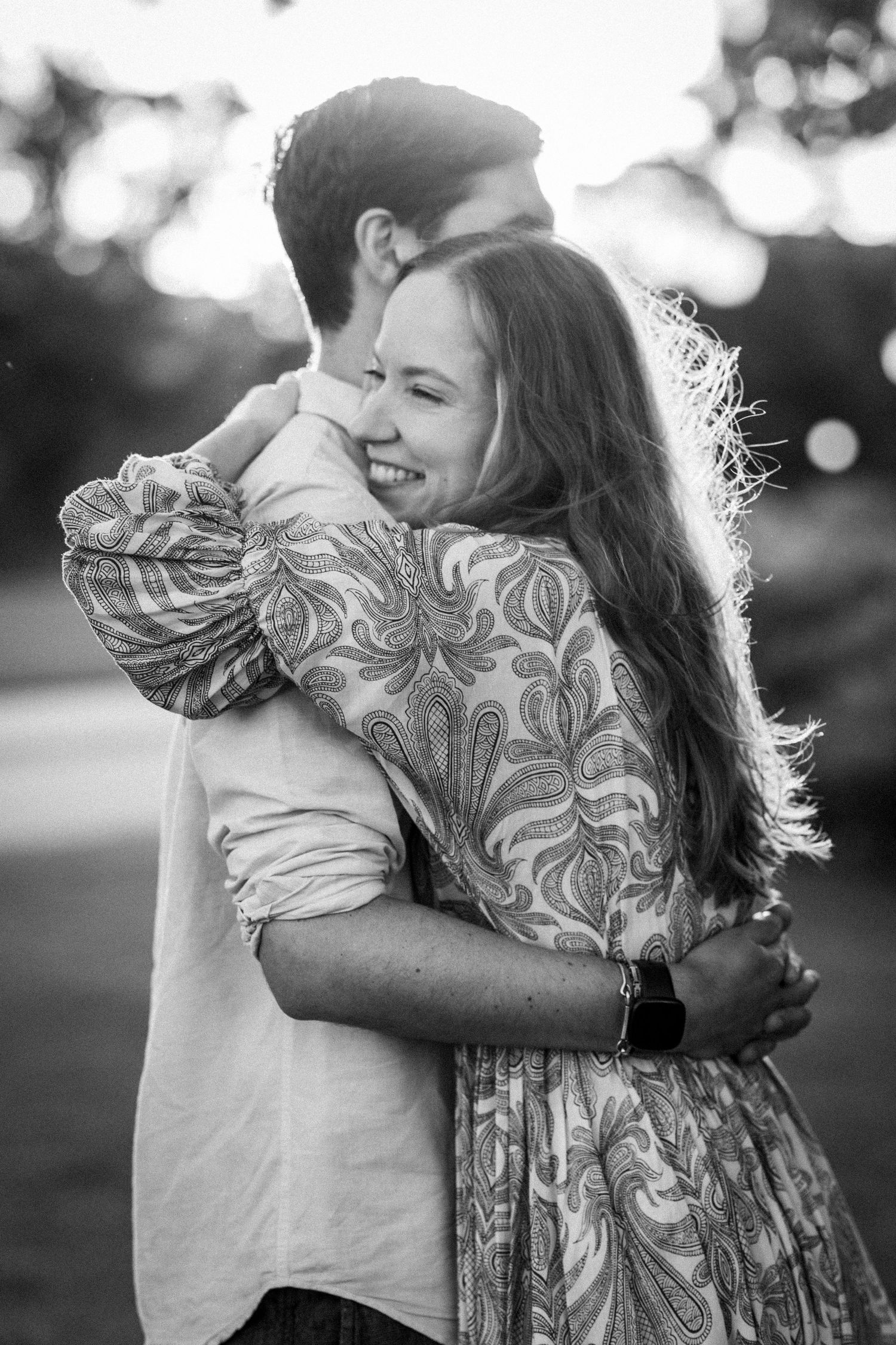 Black and white photo of a smiling couple embracing outdoors, woman in floral dress hugging man tenderly.