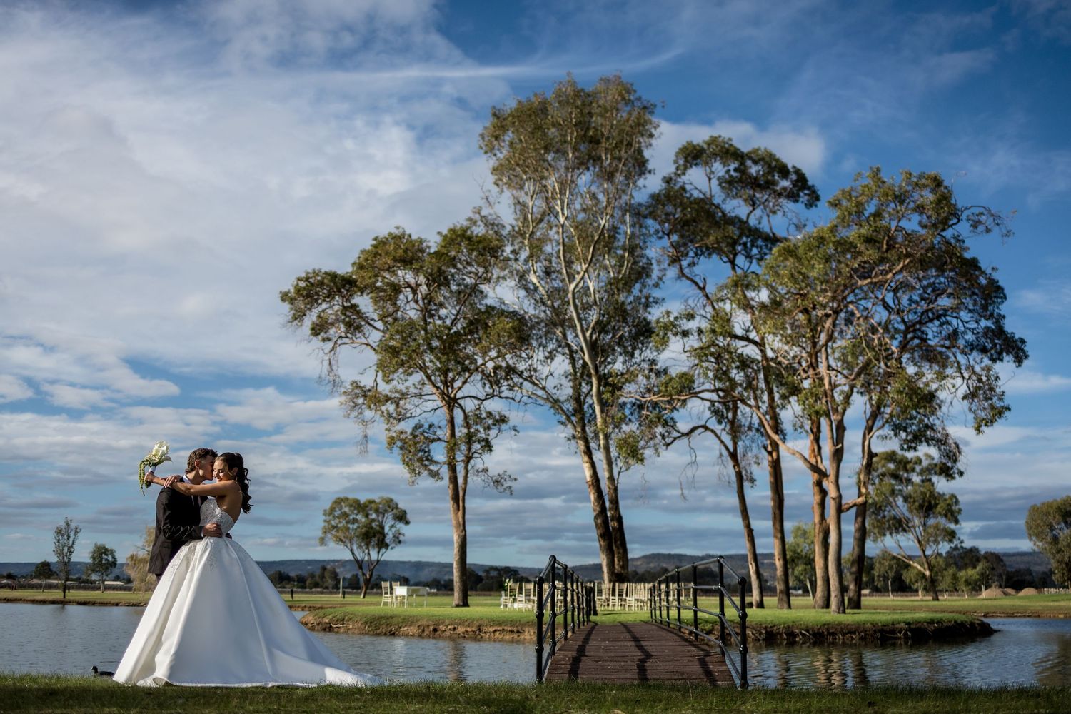 Bride and groom embrace near a serene lake surrounded by tall eucalyptus trees under a blue sky.