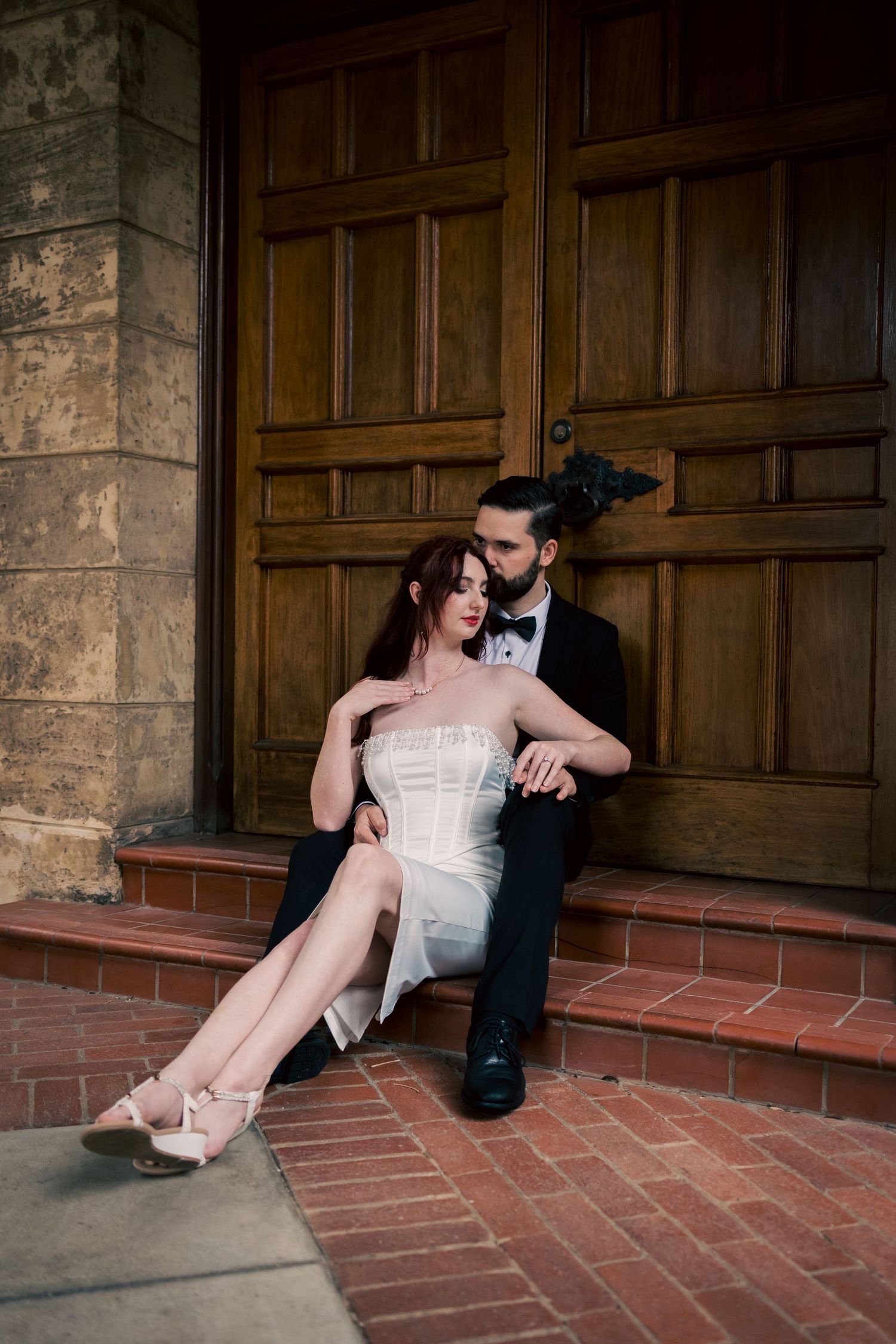 A couple in formal attire poses playfully on brick steps against wooden doors.