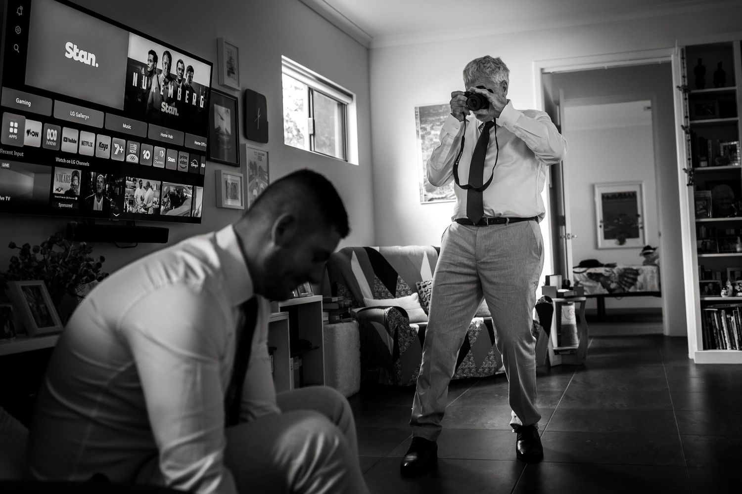 Black and white photo of a photographer capturing a man adjusting his tie in a home setting before a wedding.