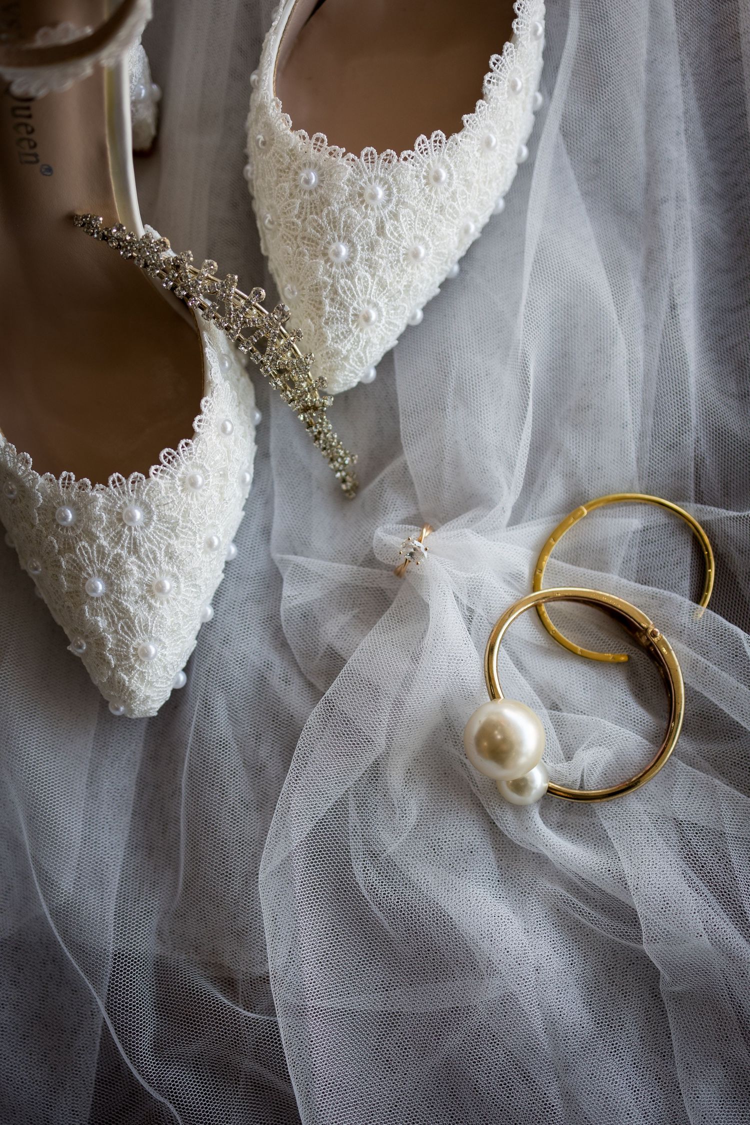 White lace bridal heels with pearl accents displayed beside gold bangles and a pearl earring on a delicate tulle veil.