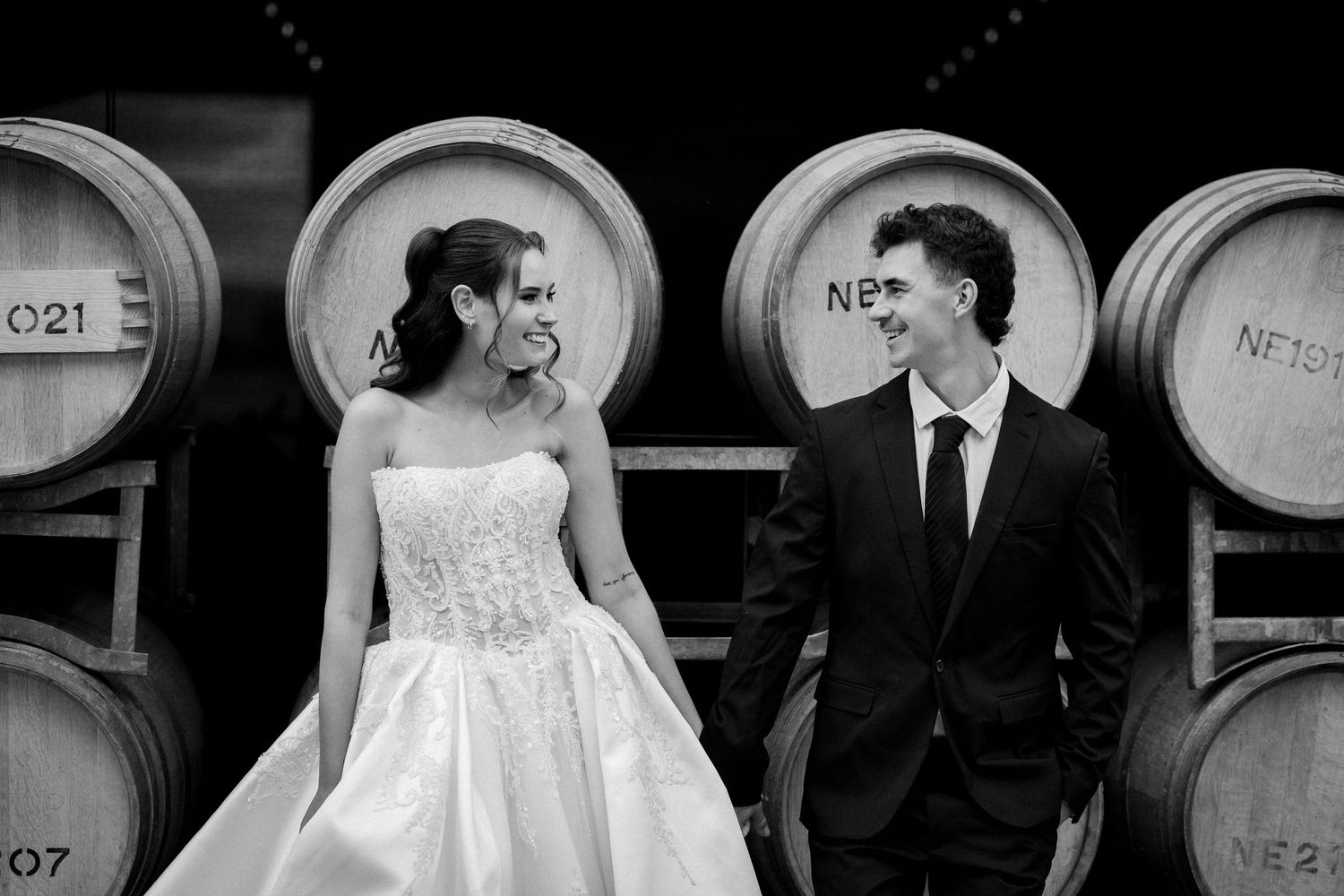 Black and white photo of a bride and groom smiling at each other in front of rustic wine barrels.