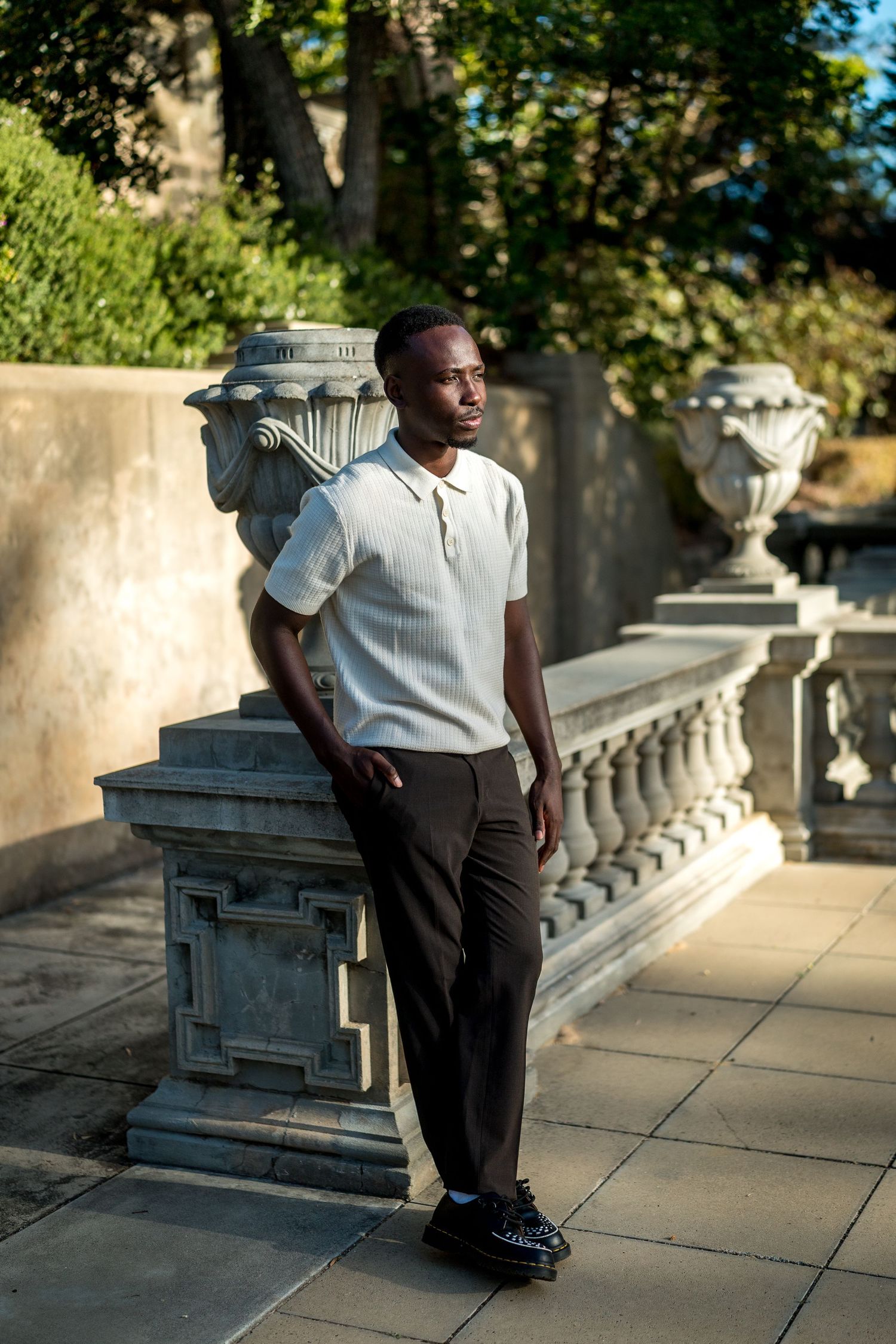 Man in a white knit polo, black trousers and black leather shoes leans against a stone balustrade beside an urn in a sunlit garden.