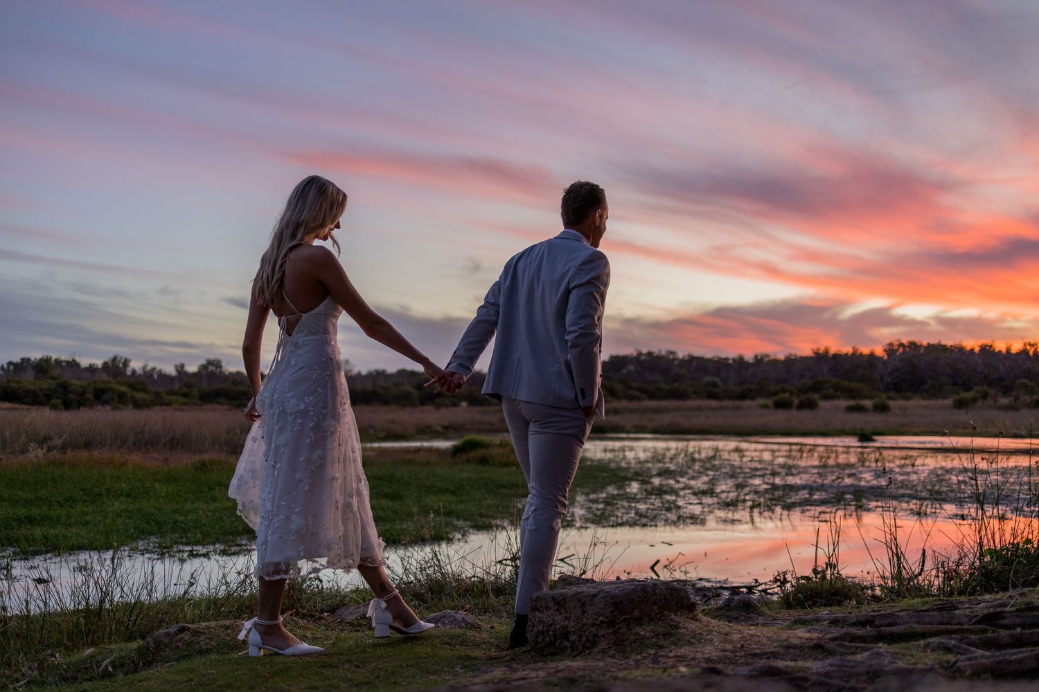 Couple holding hands at sunset by a reflective pond, bride in white lace dress, groom in gray suit.