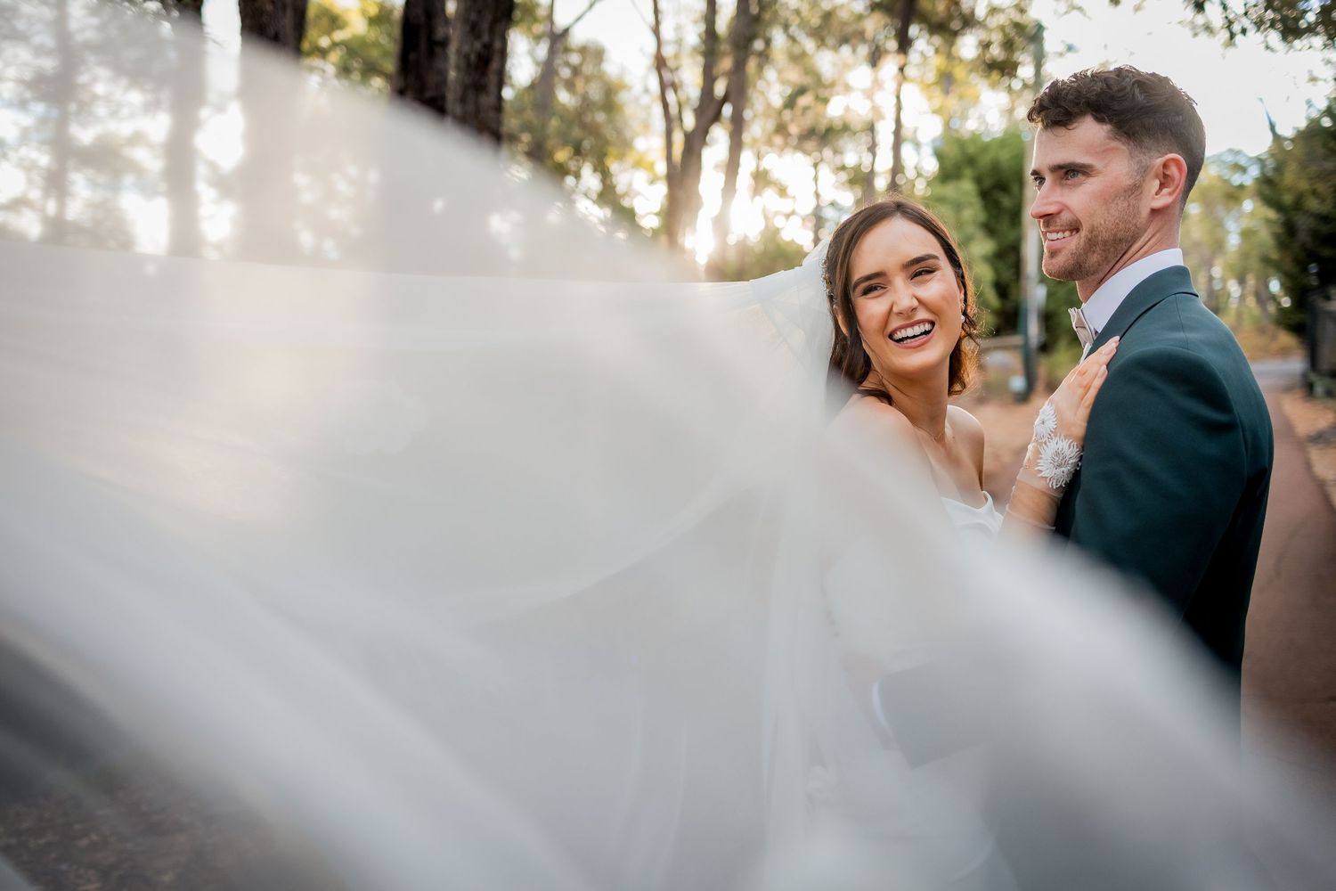 Bride and groom share a joyful moment outdoors, with her flowing white veil billowing in the breeze.