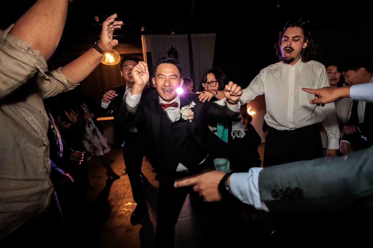 Groom in black tuxedo dancing joyfully with wedding guests at lively reception celebration.