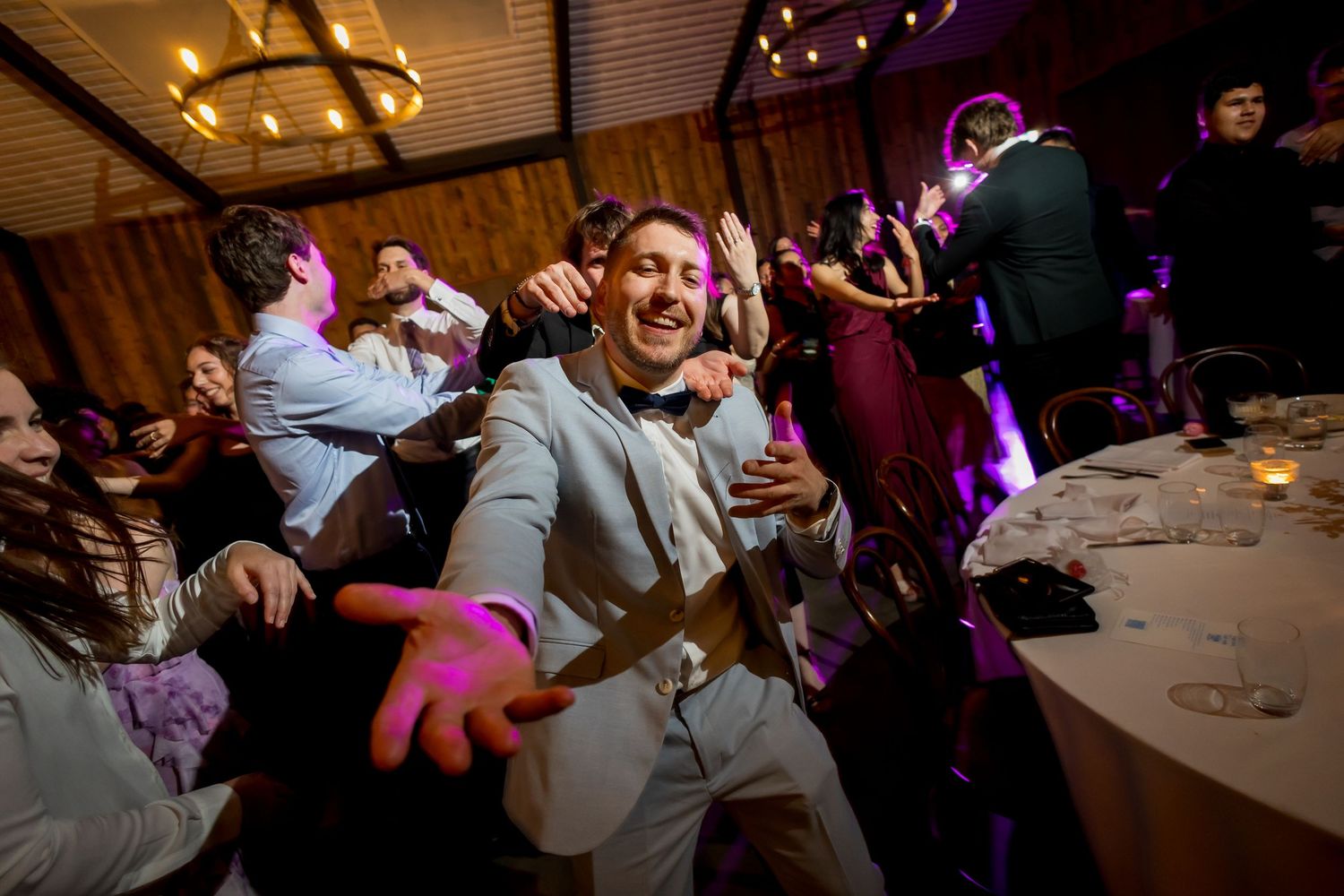 Excited groom in grey suit dances energetically at lively wedding reception with guests in background.