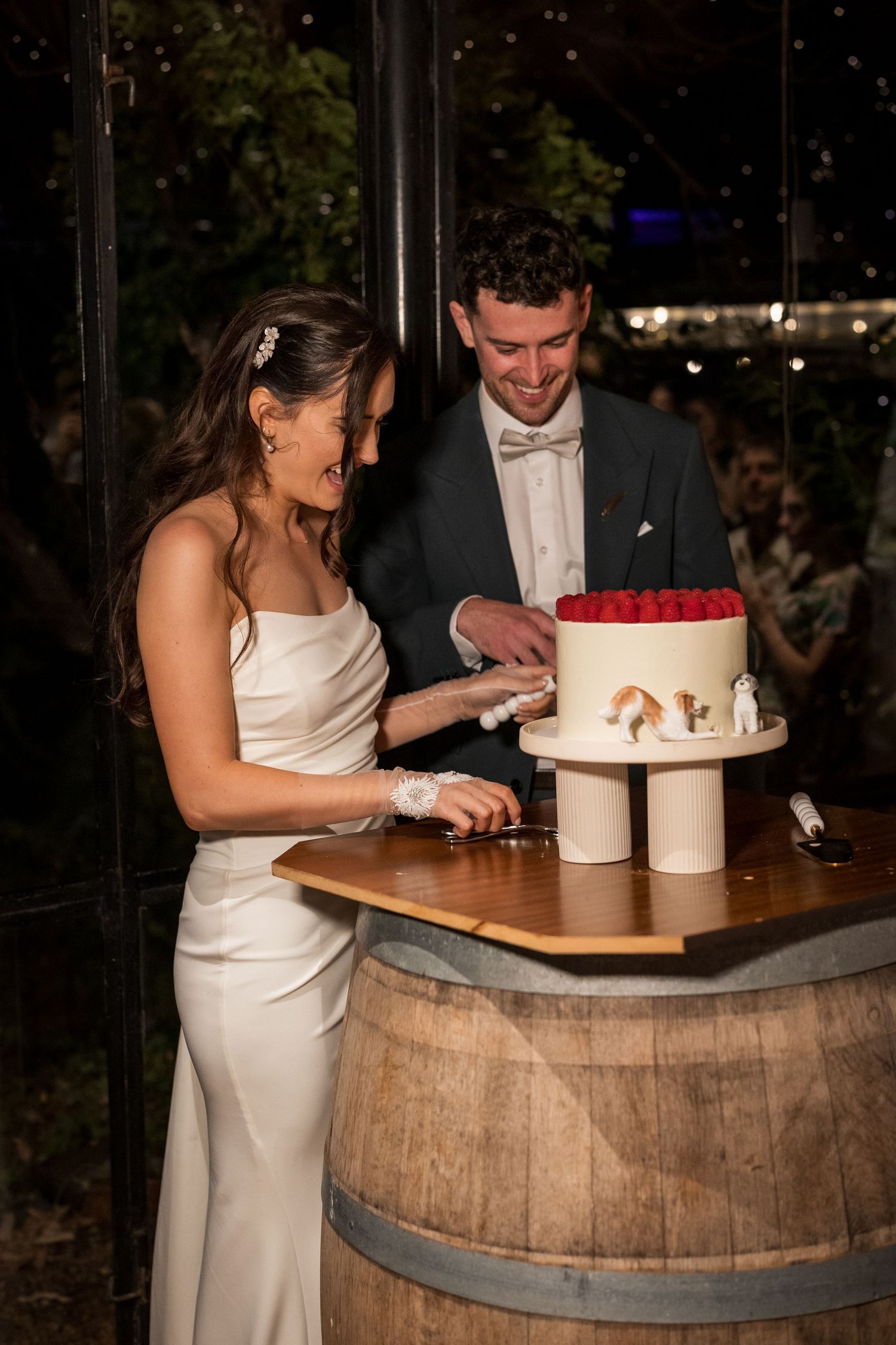 Bride and groom cut white wedding cake decorated with red roses on a wooden barrel at night.
