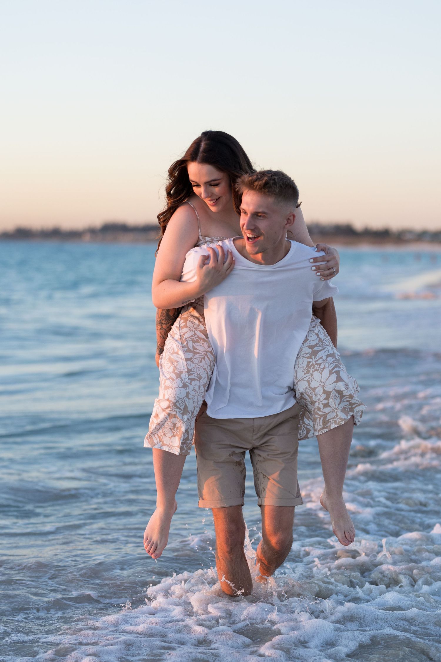 Man giving woman piggyback ride in ocean waves at sunset on beautiful sandy beach.