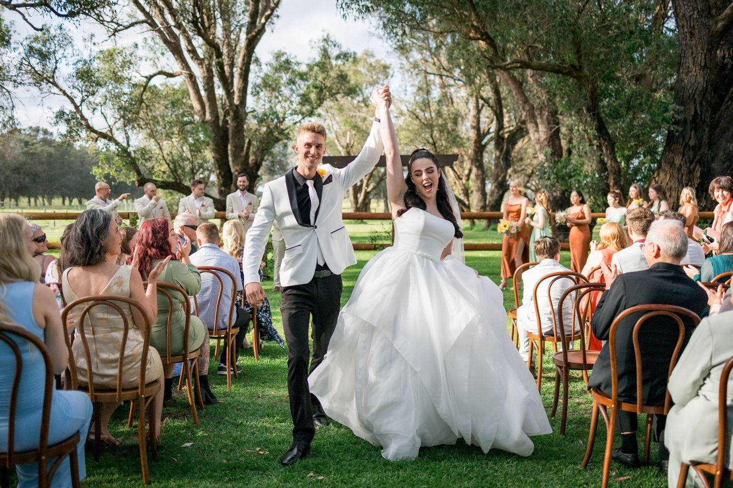 Joyful newlyweds raise hands in celebration as they walk down the aisle at an outdoor garden wedding ceremony.