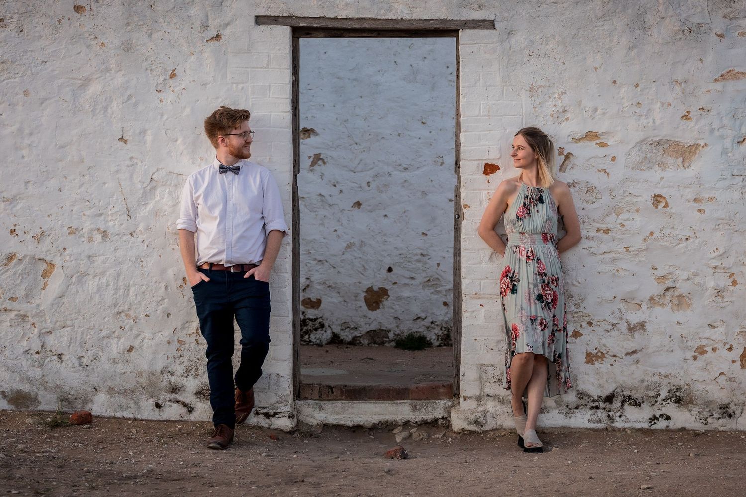 Couple in casual attire poses against rustic white wall with doorway.