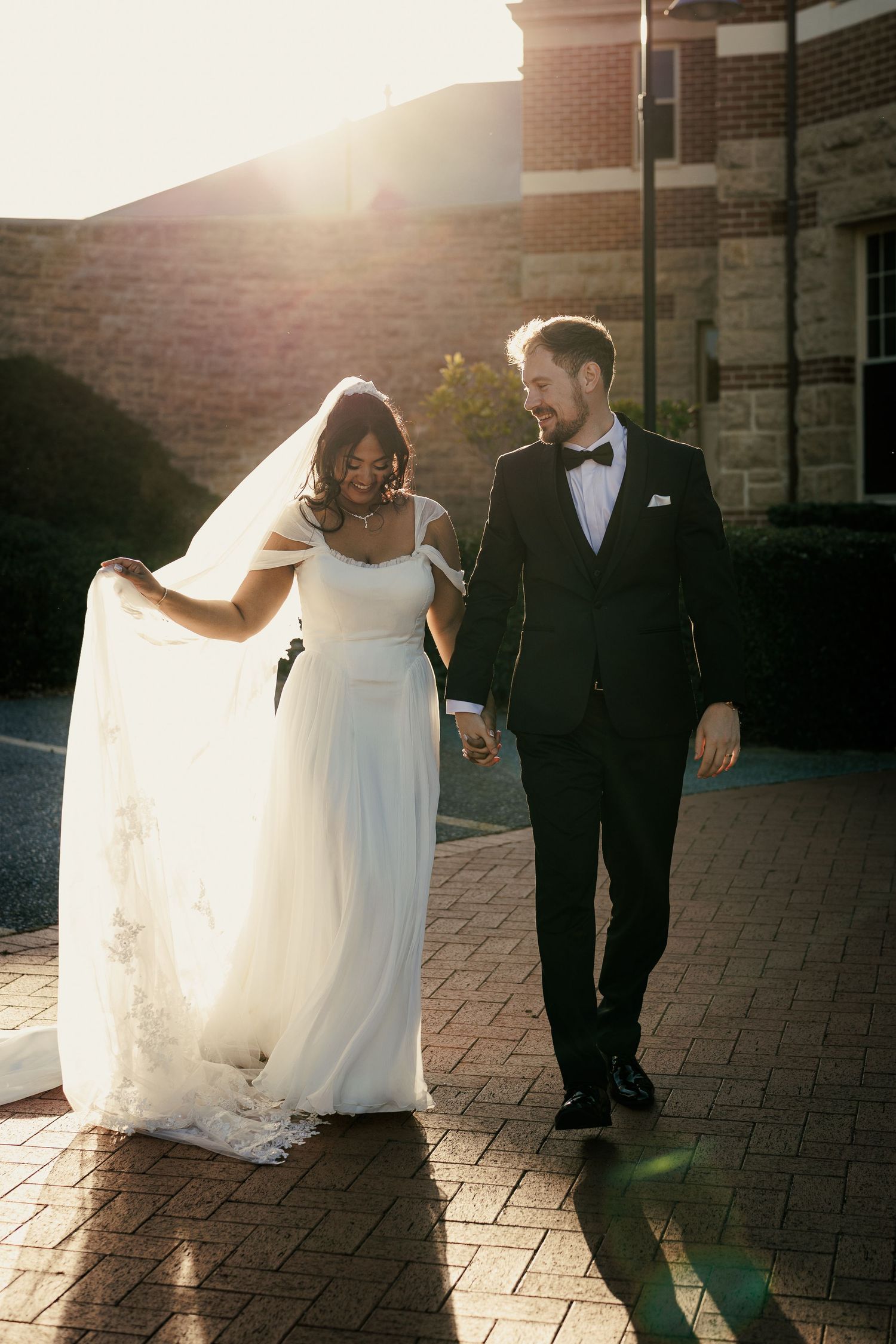 A bride and groom walk hand-in-hand on a brick path, bathed in golden sunset light, as she lifts her flowing white veil.