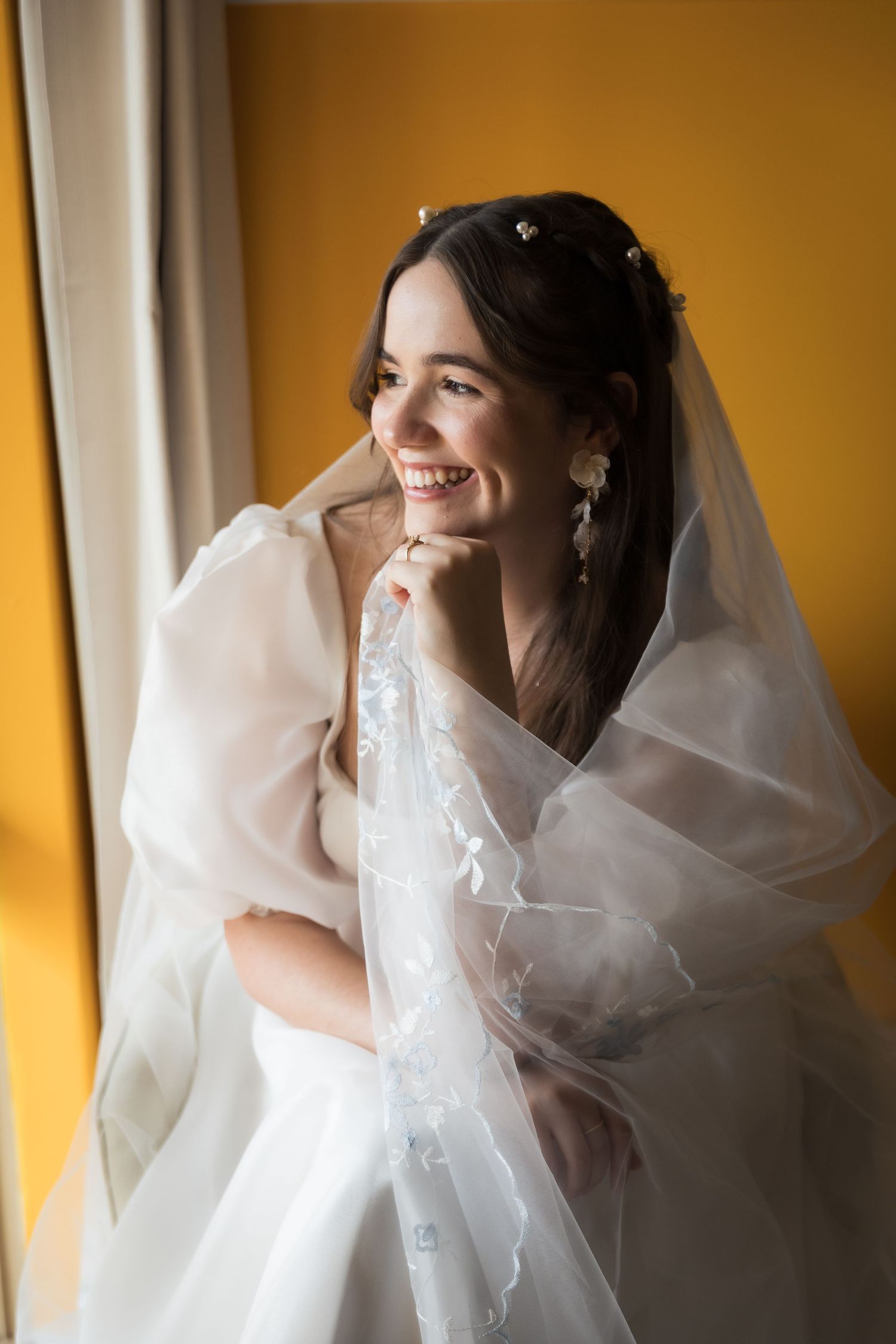 Smiling bride in white veil and gown poses by window with yellow wall on her wedding day.