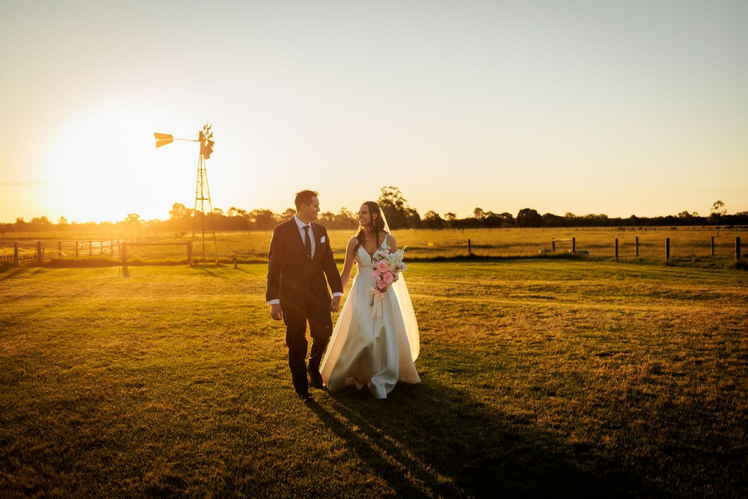 Bride and groom walk hand in hand across a golden sunlit farm field at sunset with a windmill in the background.