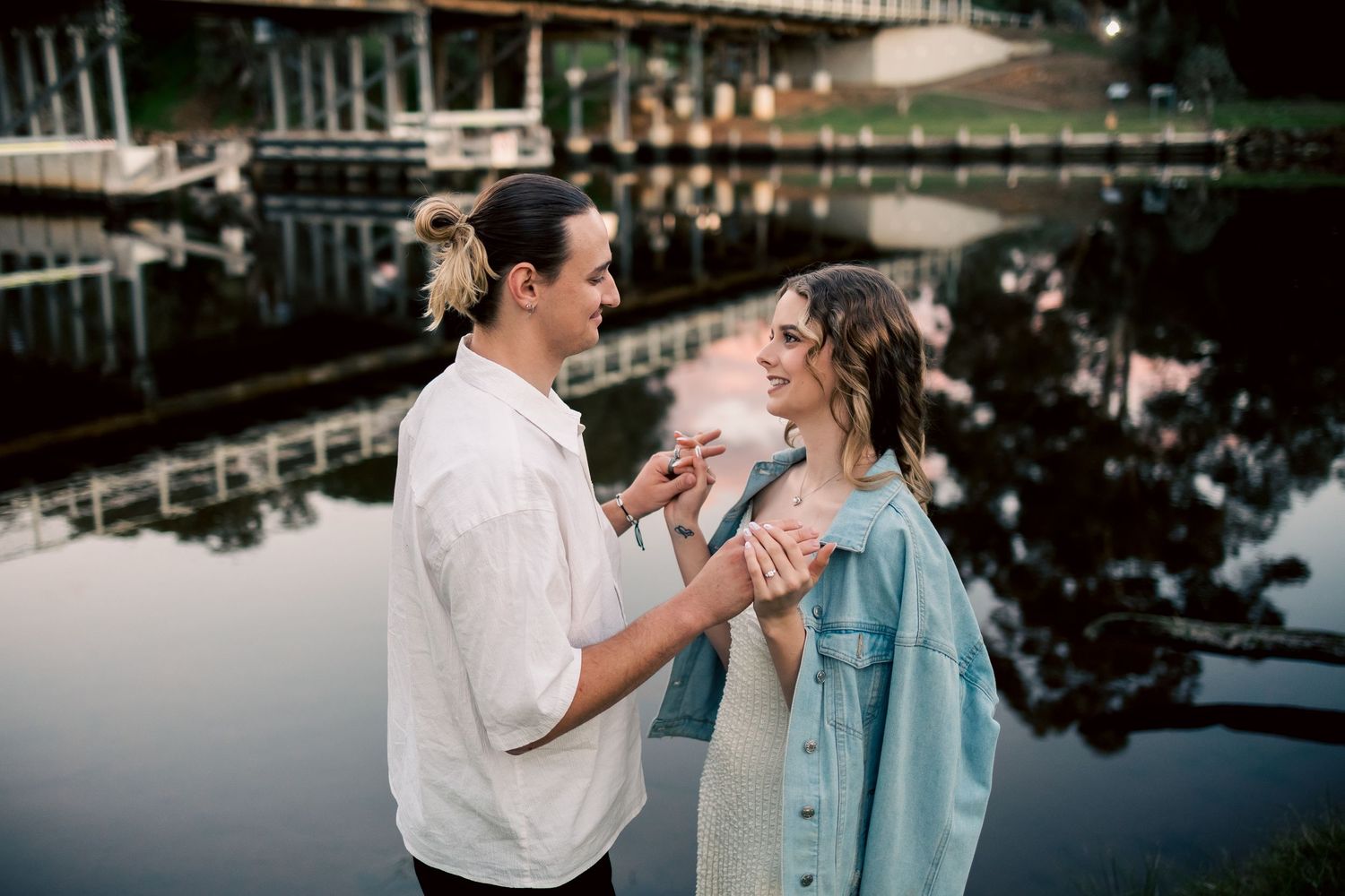 A couple holds hands and smiles at each other near a reflective river with a bridge in the background at dusk at Guildford, Perth