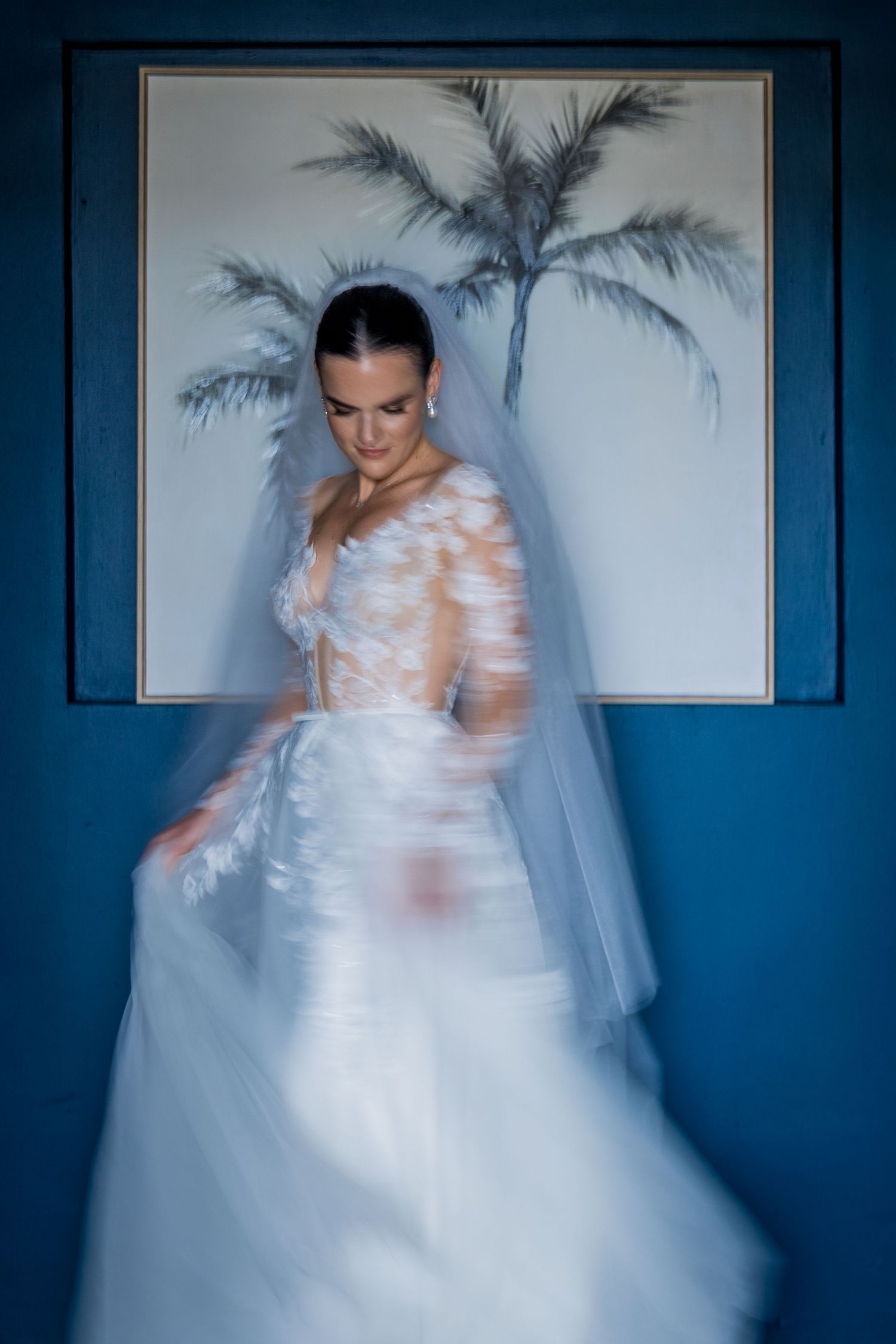 Bride in lace wedding gown and veil twirling before blue wall with palm tree art.